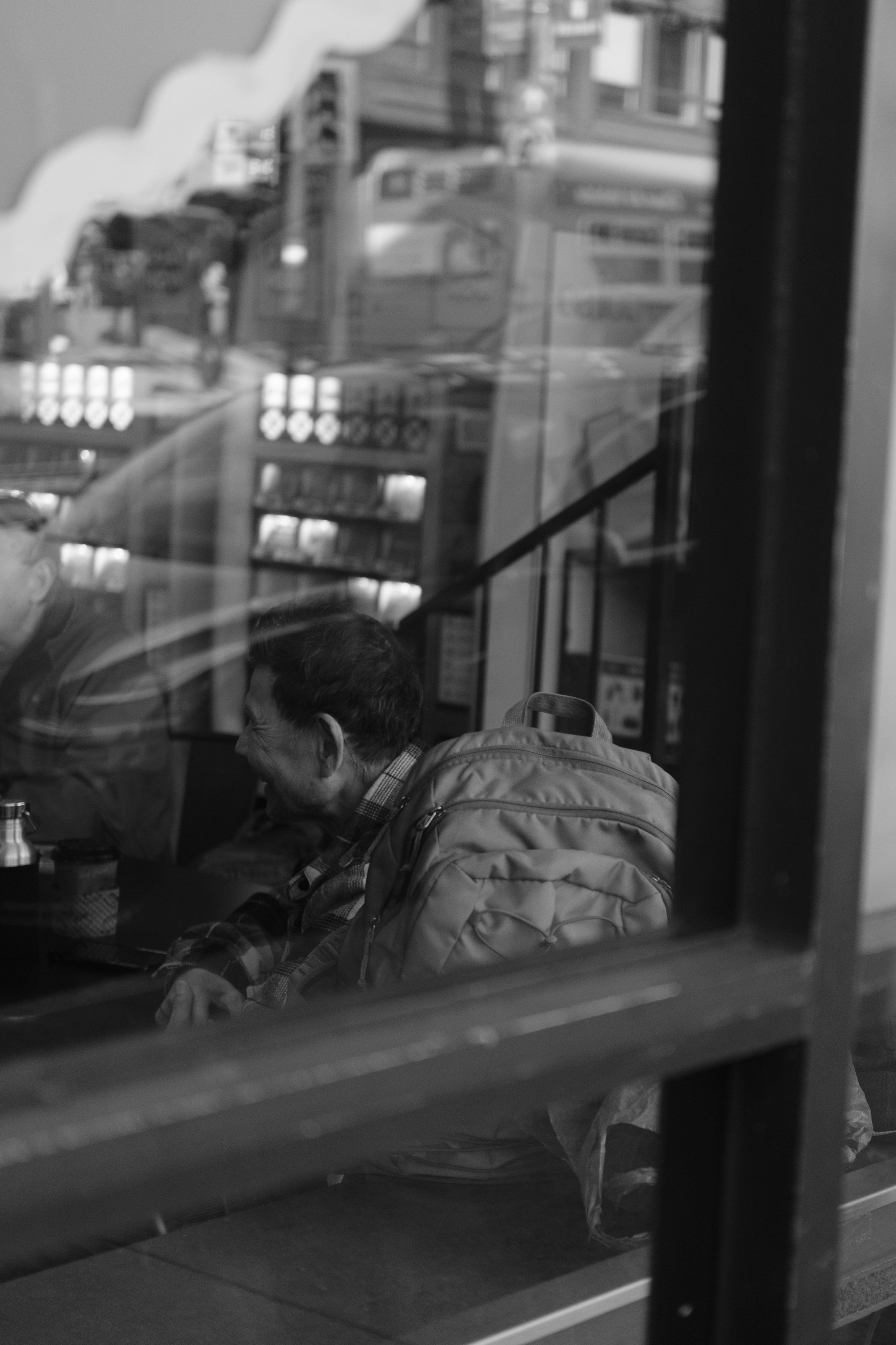 A man with a backpack sitting inside a cafe, viewed through a window with reflections of the street outside.