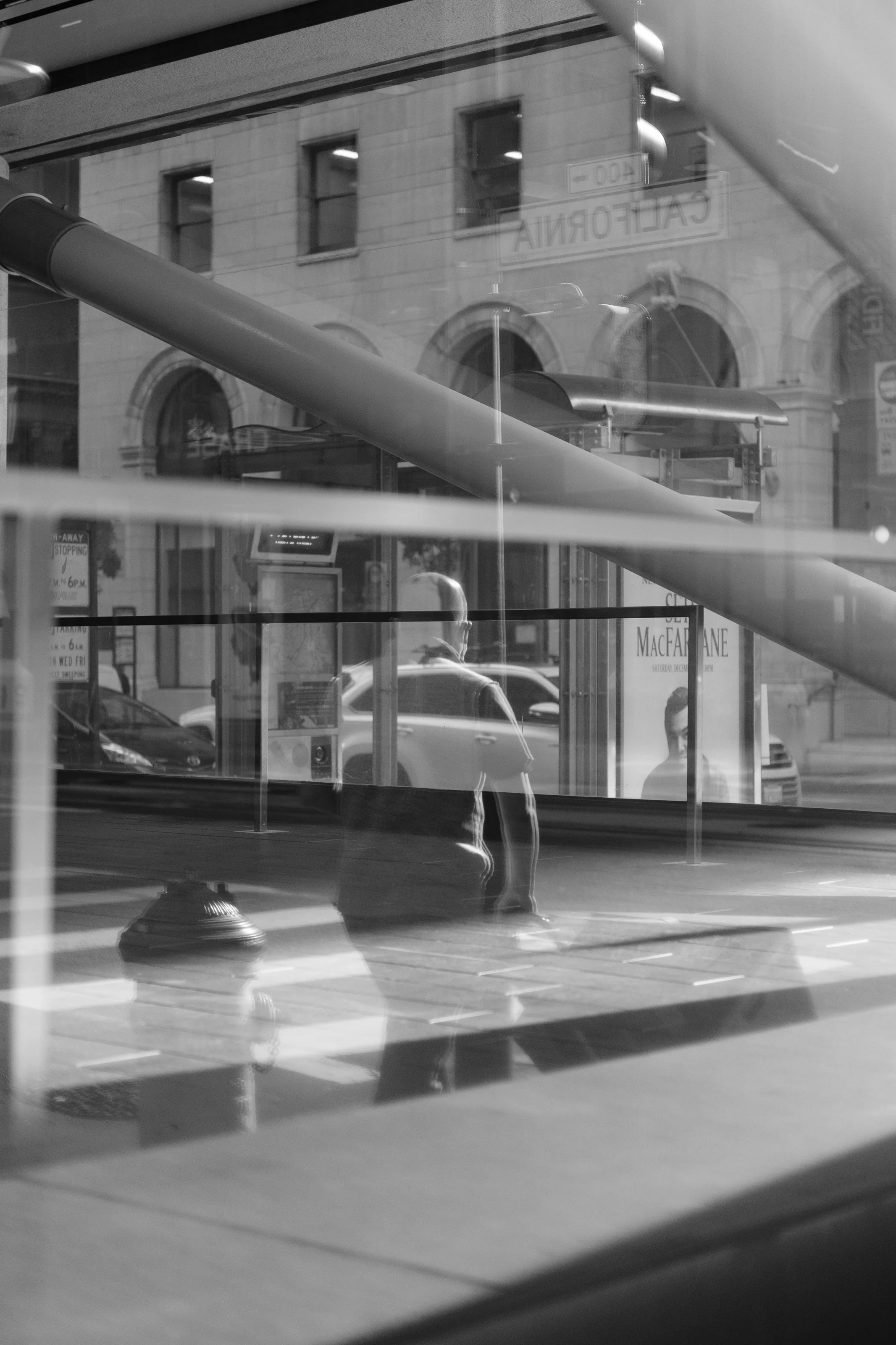 A black-and-white photo of a city street seen through a window, with reflections of a man walking, buildings, cars, and signs visible.