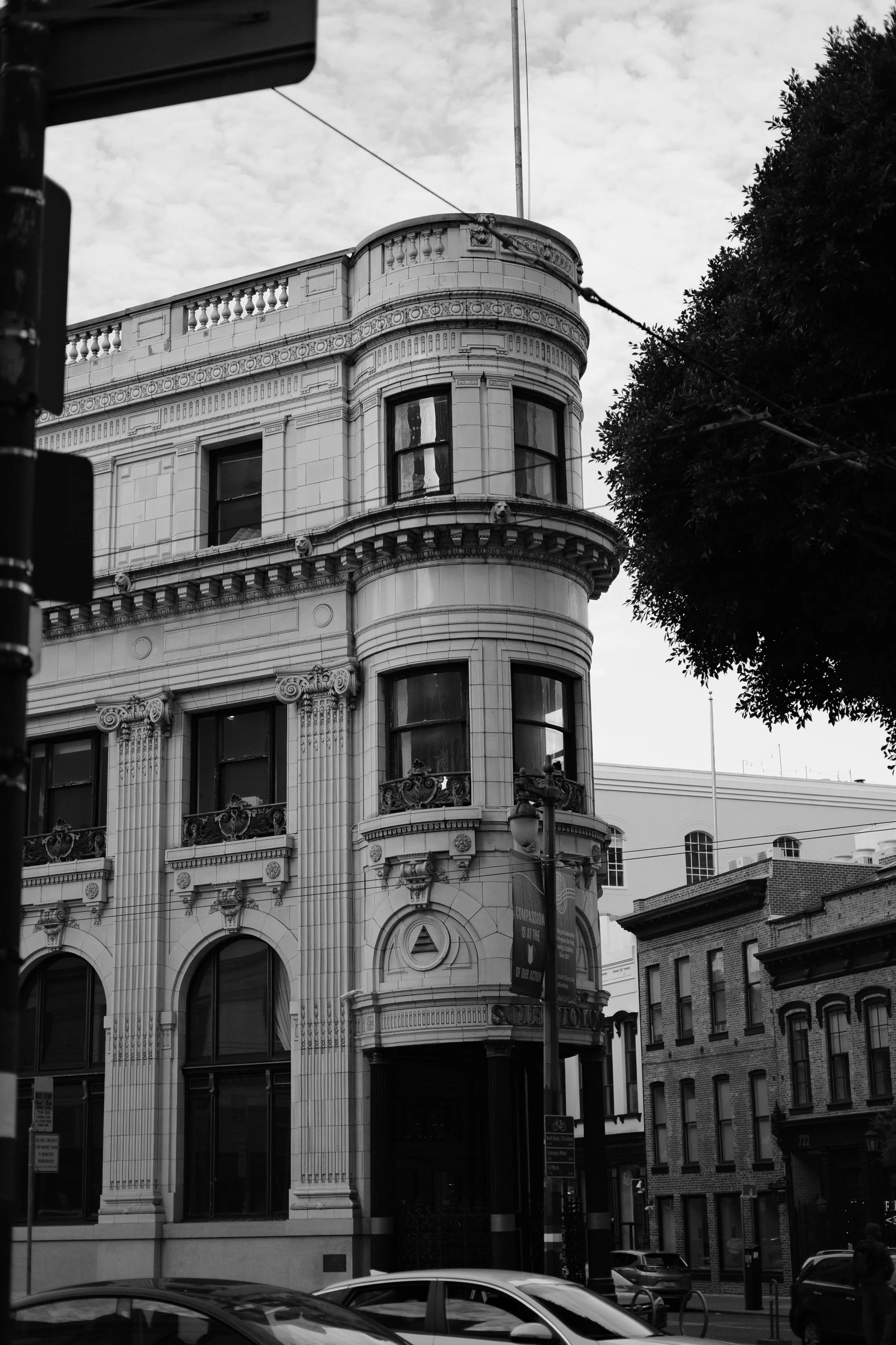 Black and white photo of an ornate, historic building with rounded corners, decorative columns, and large windows, located on a city street with cars parked in front and a leafy tree on the right.