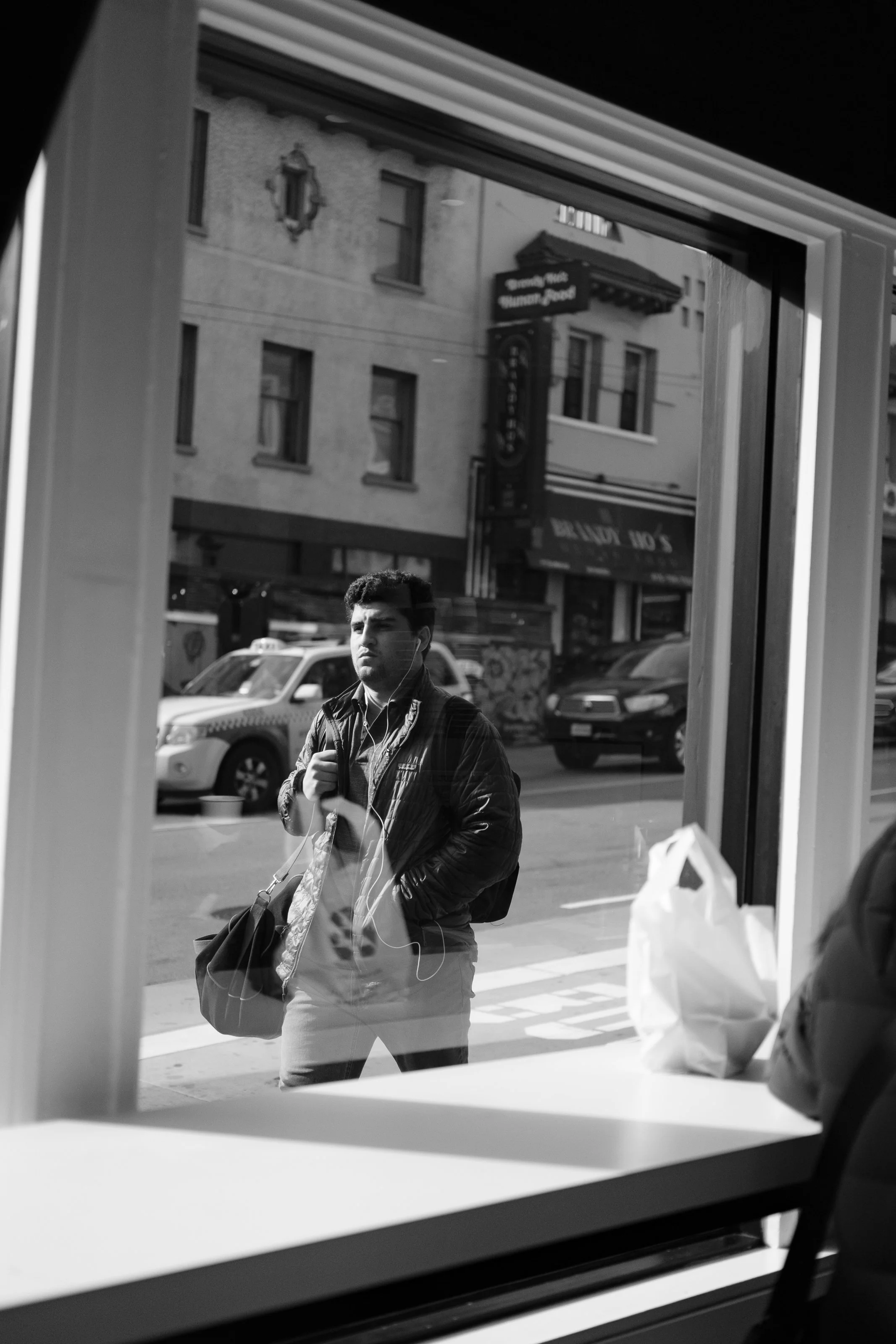 View of a street scene through a window, with a man standing outside holding a bag and wearing earphones, reflected in the glass, with buildings and cars in the background.