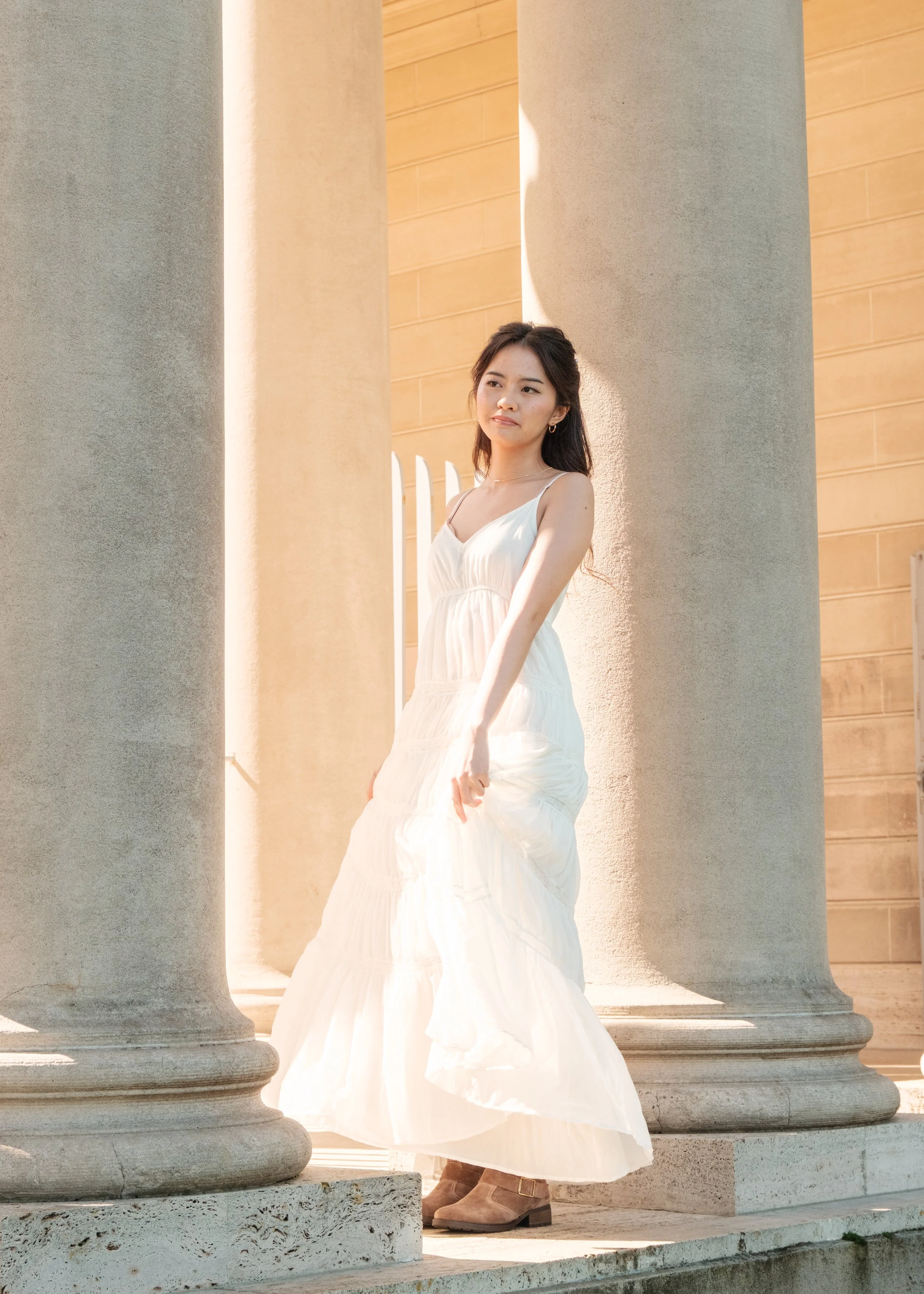A woman in a white summer dress standing between large stone columns outside a building with brick walls, sunlight illuminating her face.
