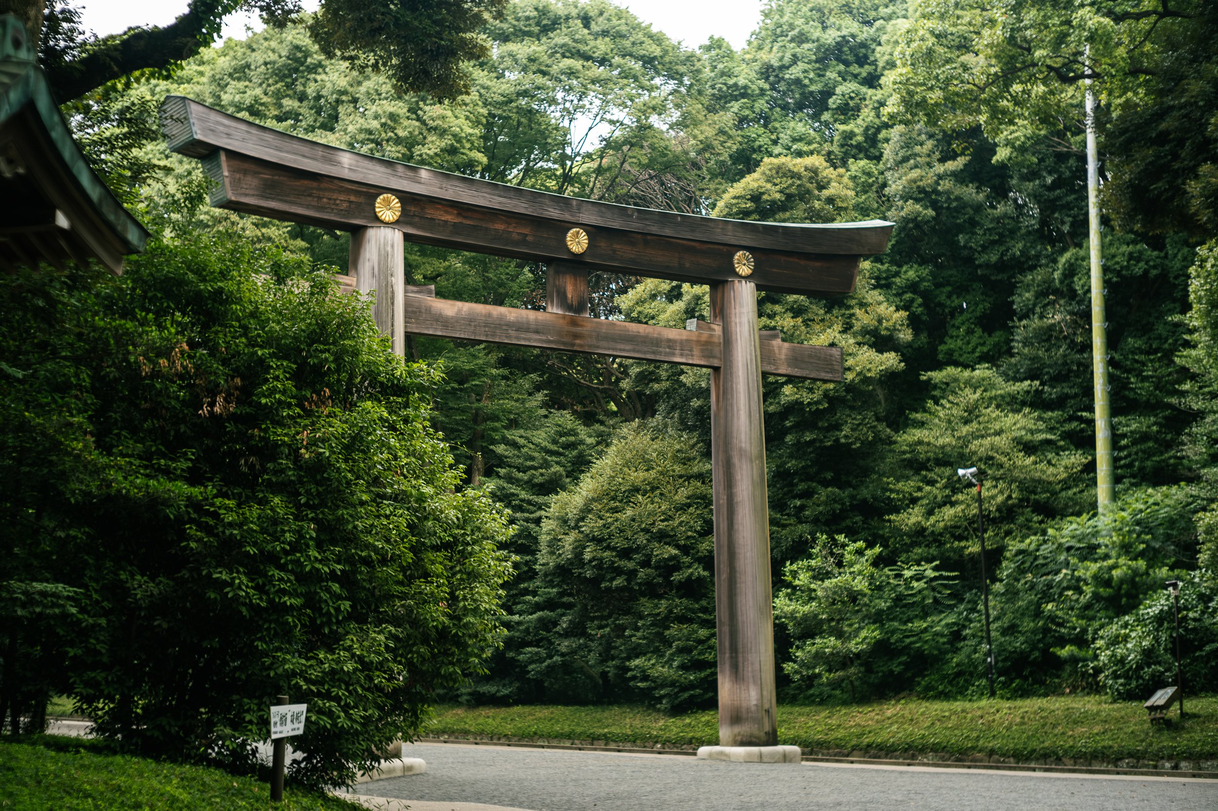 A large traditional Japanese torii gate made of wood with three gold embellishments, standing amidst lush green trees and shrubbery.