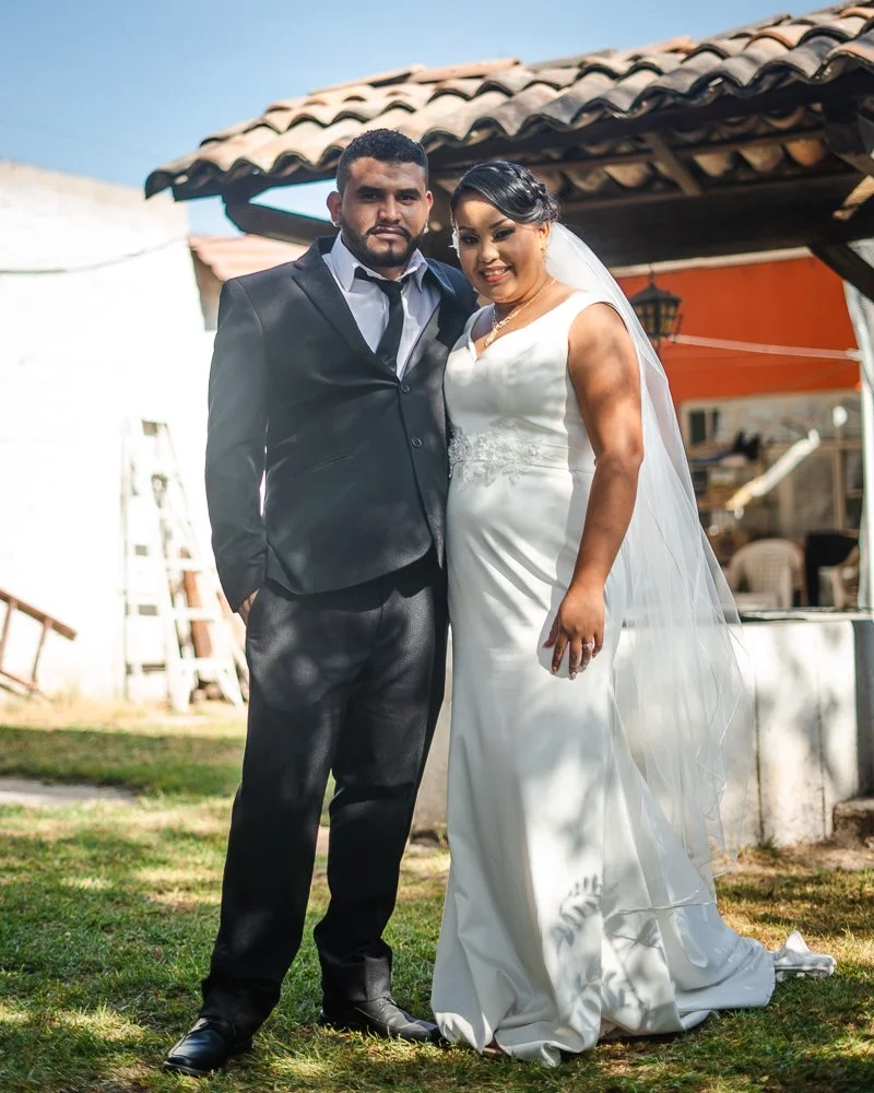 A bride and groom standing outdoors on their wedding day, with the bride in a white wedding gown and veil, and the groom in a black suit with a white shirt, against a backdrop of a rustic building and blue sky.