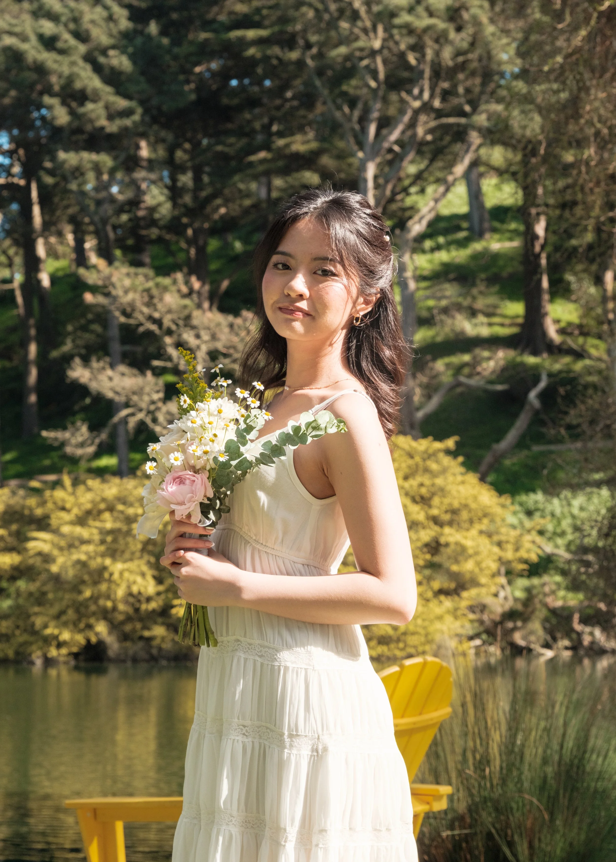 Young woman standing outdoors holding a bouquet of white and pink flowers with greenery, in front of a lake and surrounded by trees and greenery.