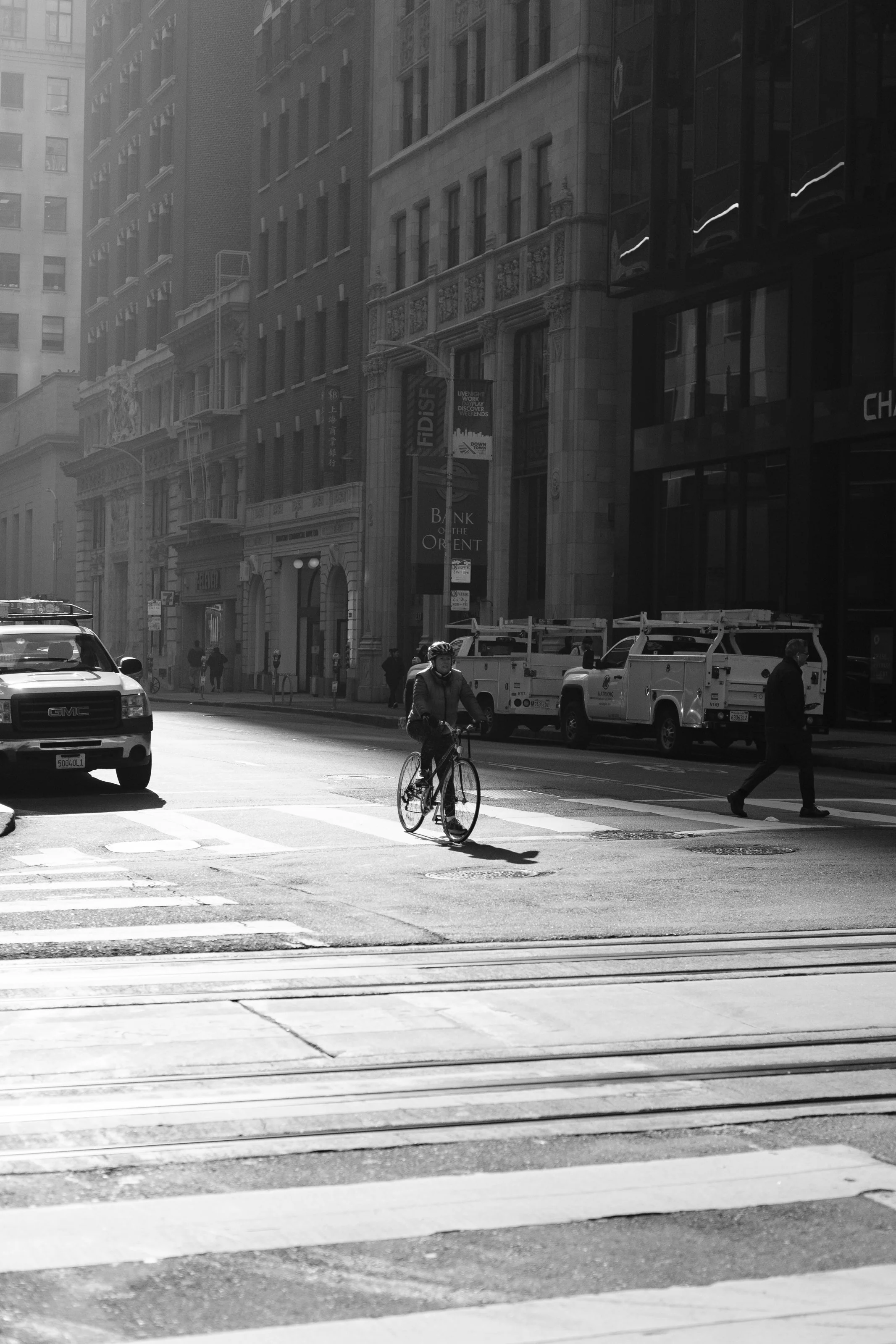 A black and white photo of a city street with a cyclist crossing at a crosswalk, parked cars, and pedestrians, with tall buildings in the background.
