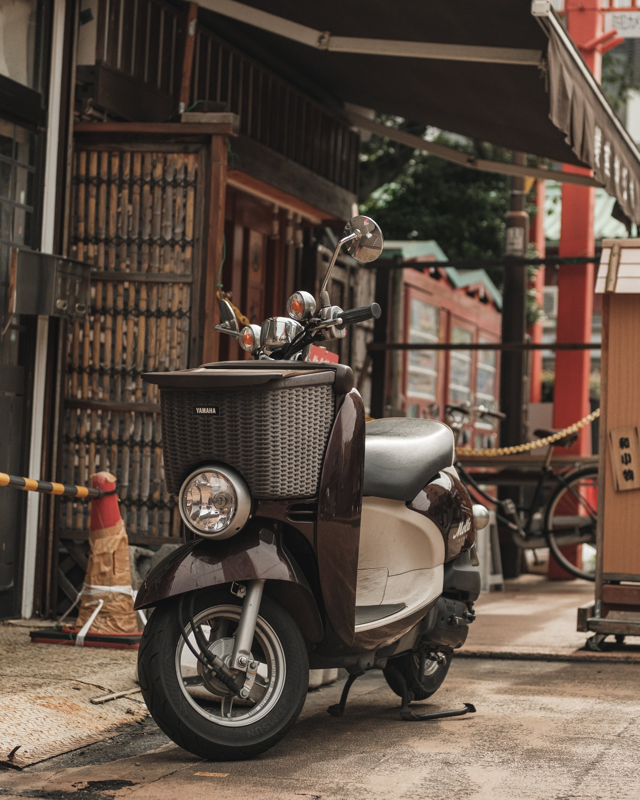 A Yamaha scooter parked on the street in front of a traditional building with wooden elements and a red torii gate in the background.