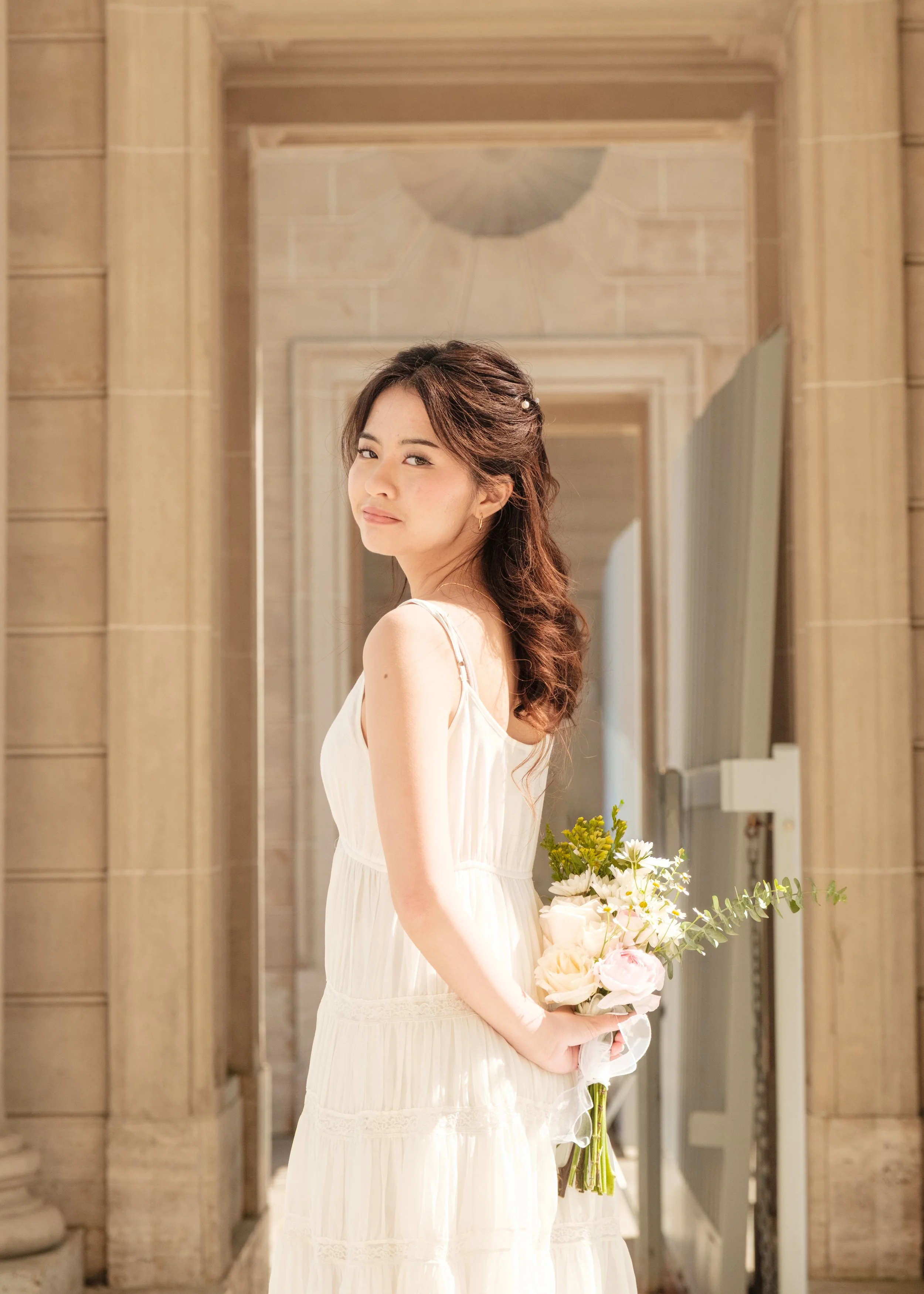 A woman with wavy brown hair holding a bouquet of white and pink flowers, wearing a white dress, standing in a brightly lit, ornate hallway with beige stone walls and arched doorway.
