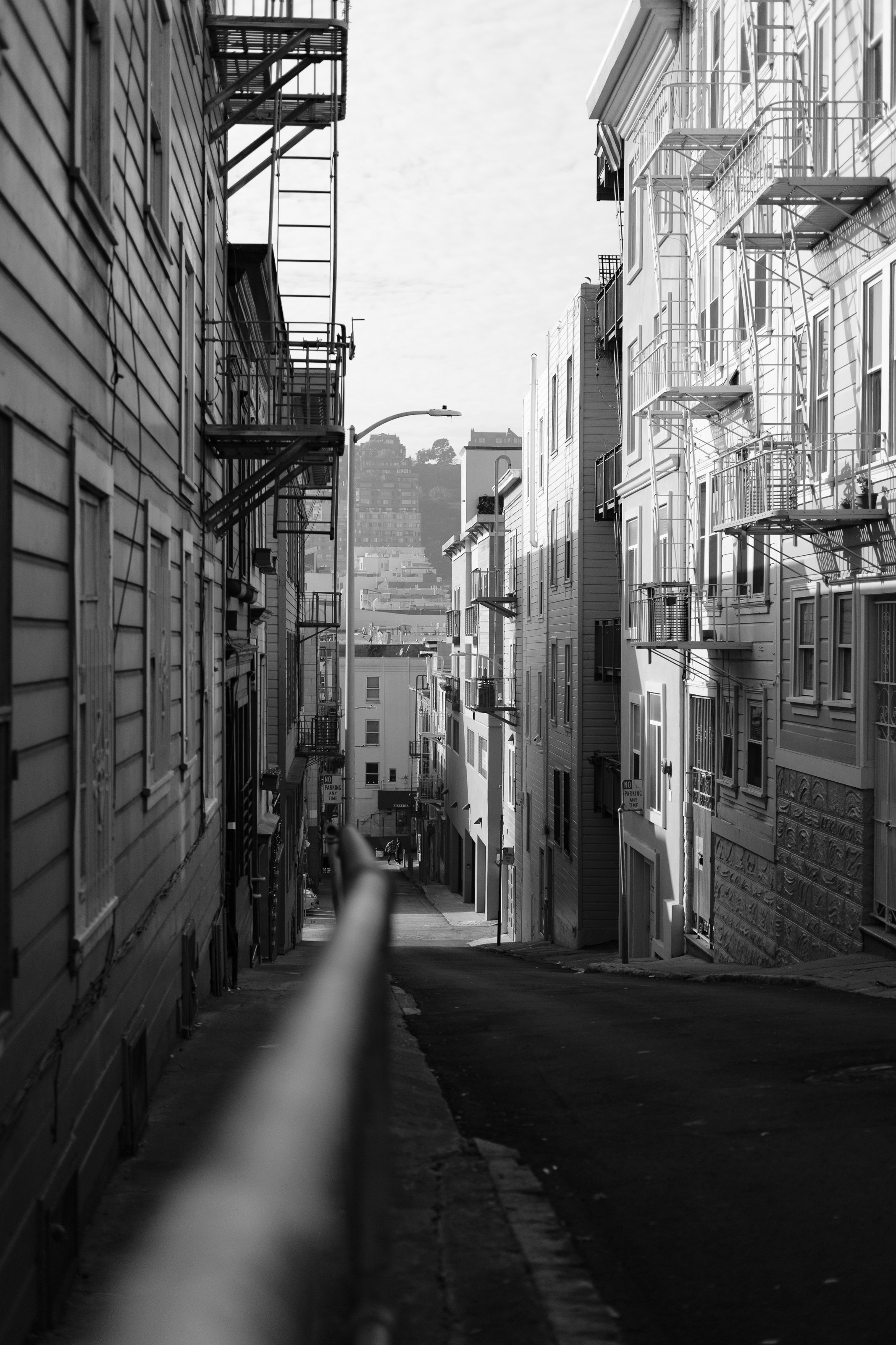 Black and white photo of a steep city street flanked by tall buildings with fire escapes, leading to a hill in the distance, with a lamppost and a blurred handrail in the foreground.