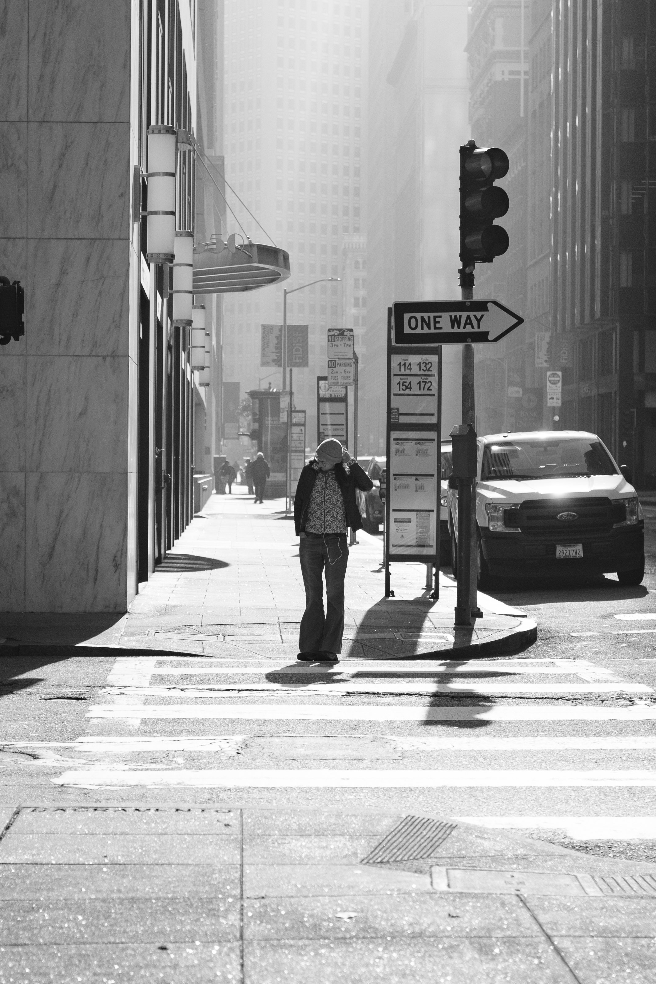A black and white photo of a city street scene with a woman standing on a crosswalk, talking on her cellphone, with tall buildings and cars in the background.