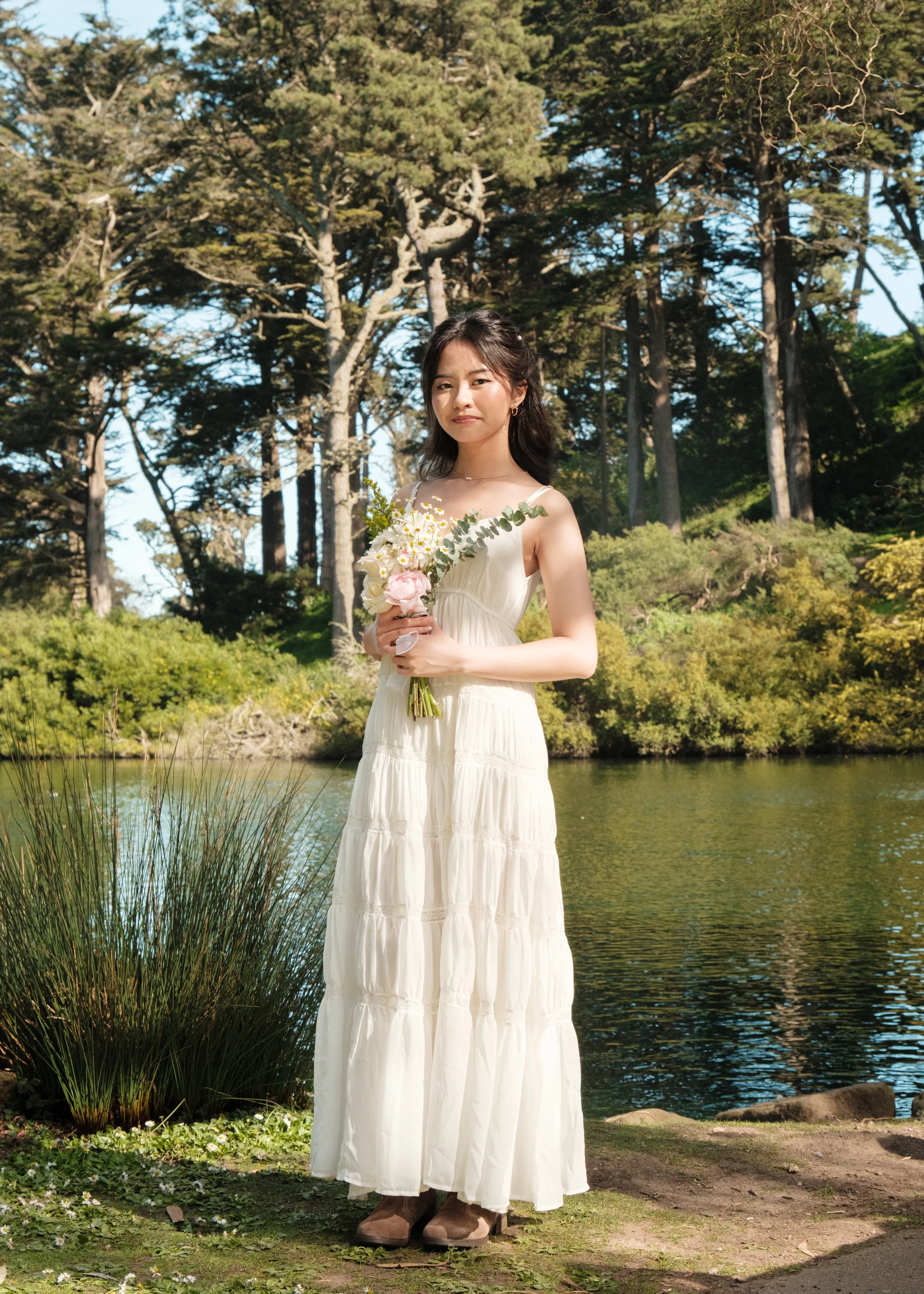 A young woman in a white sleeveless dress holds a floral bouquet, standing near a pond surrounded by trees and greenery on a sunny day.