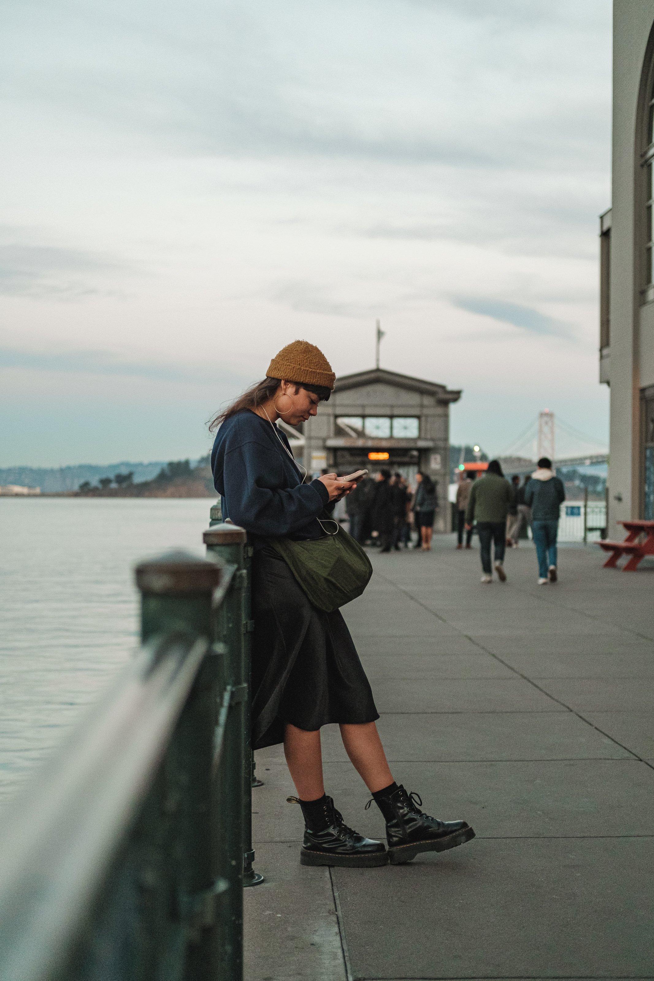 A woman standing on a waterfront pier, looking at her phone, wearing a brown knit hat, dark clothing, and black combat boots, with a group of people and a bridge in the background.