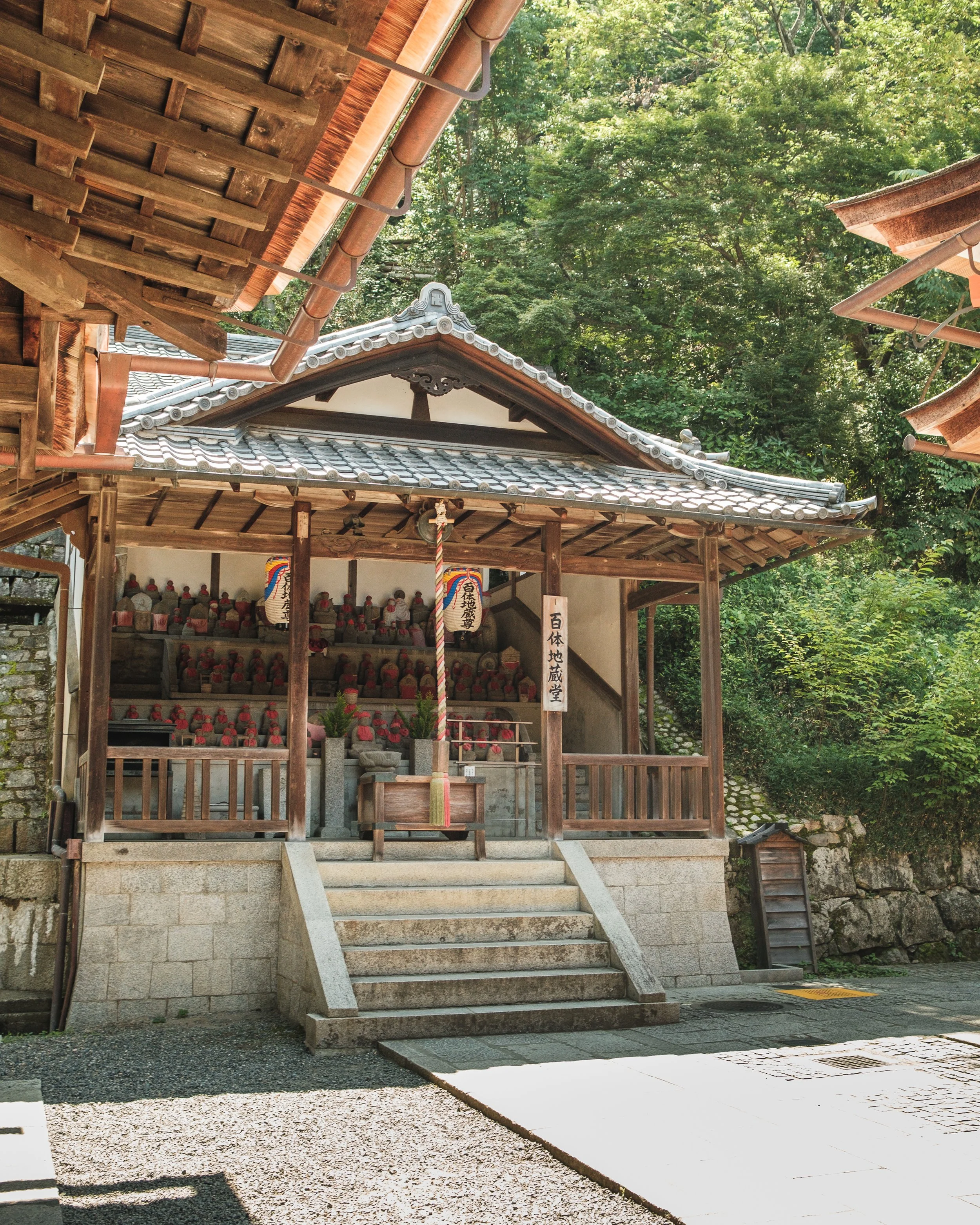 A small traditional shrine surrounded by green trees, with steps leading up to it and many small figurines inside.