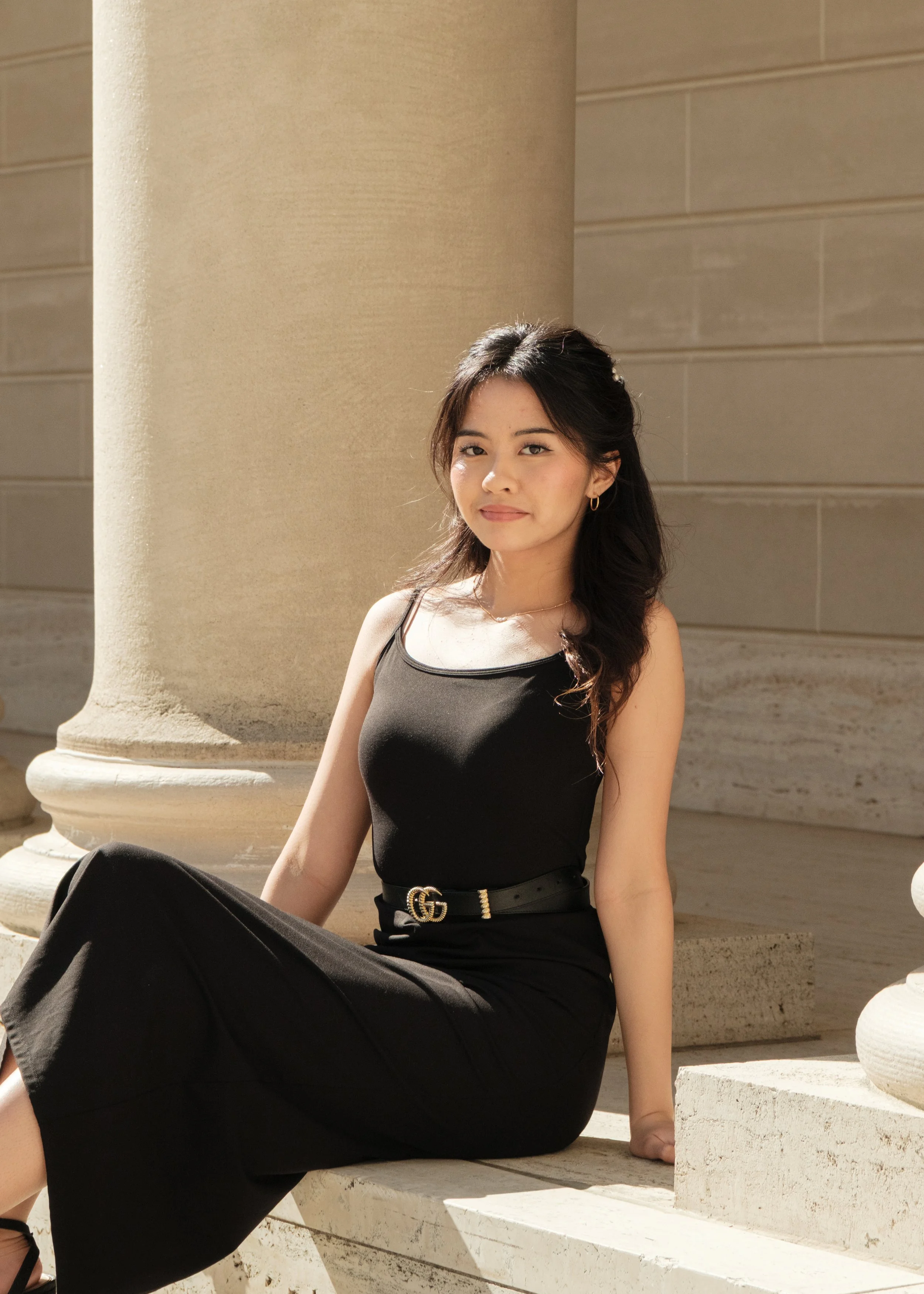 A young woman sitting outdoors near a large stone column, wearing a black dress and a Gucci belt, with long wavy dark hair, posing for a photo in sunlight.