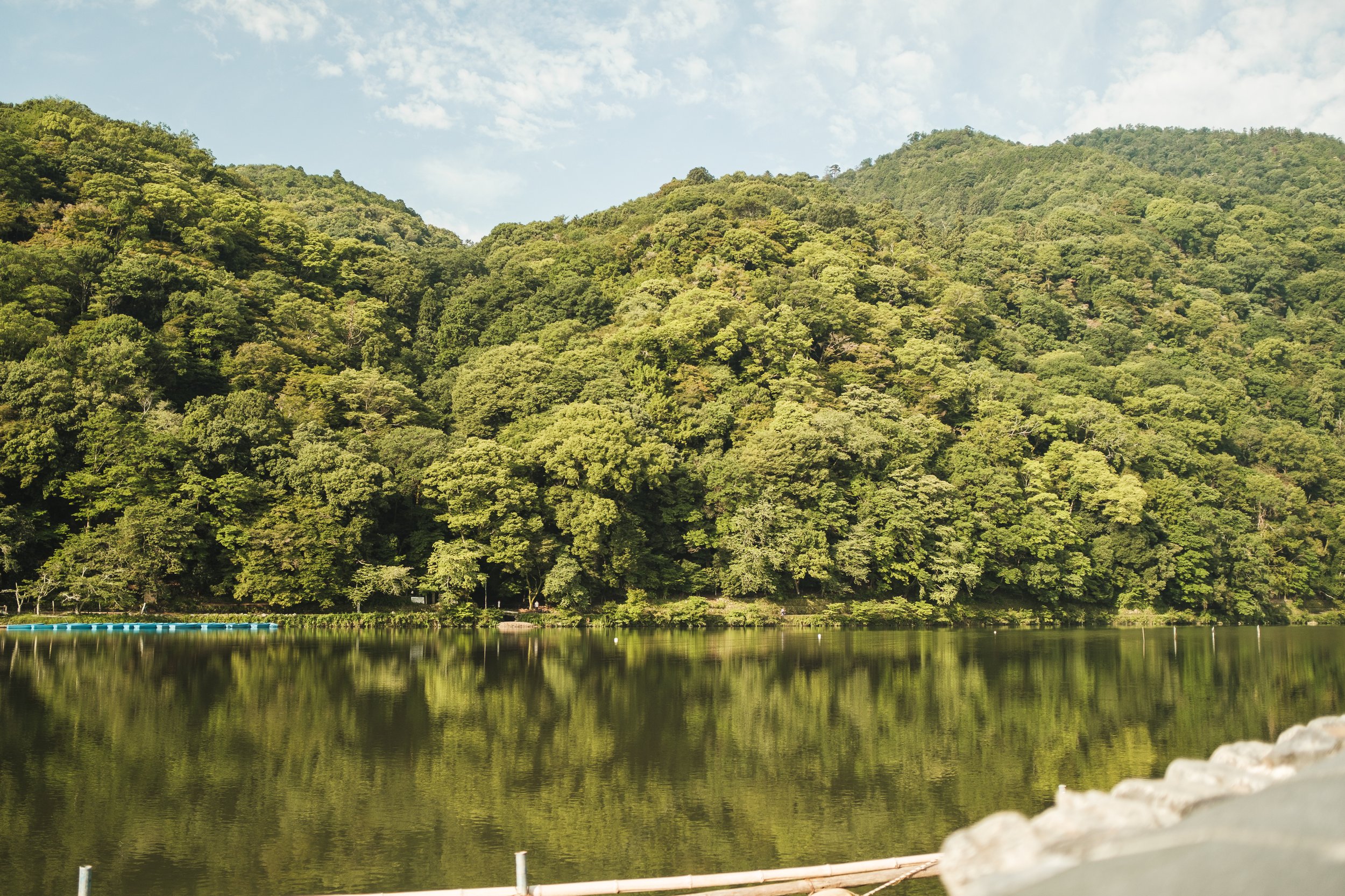 A calm lake reflecting green trees on a hillside with blue sky and scattered white clouds.