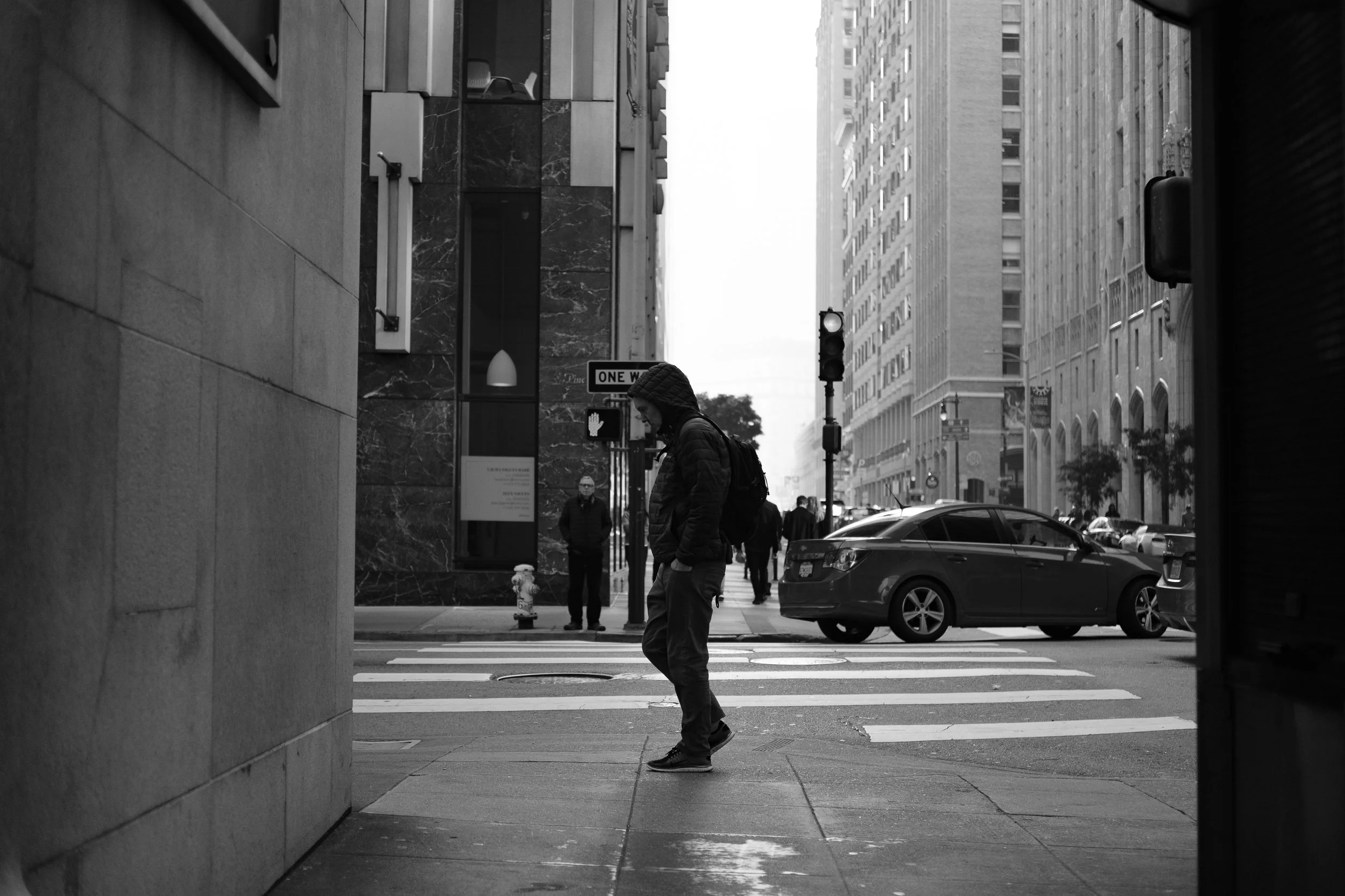 A man in a hoodie and backpack walking on a city sidewalk, with people and cars in the background, in an urban setting.