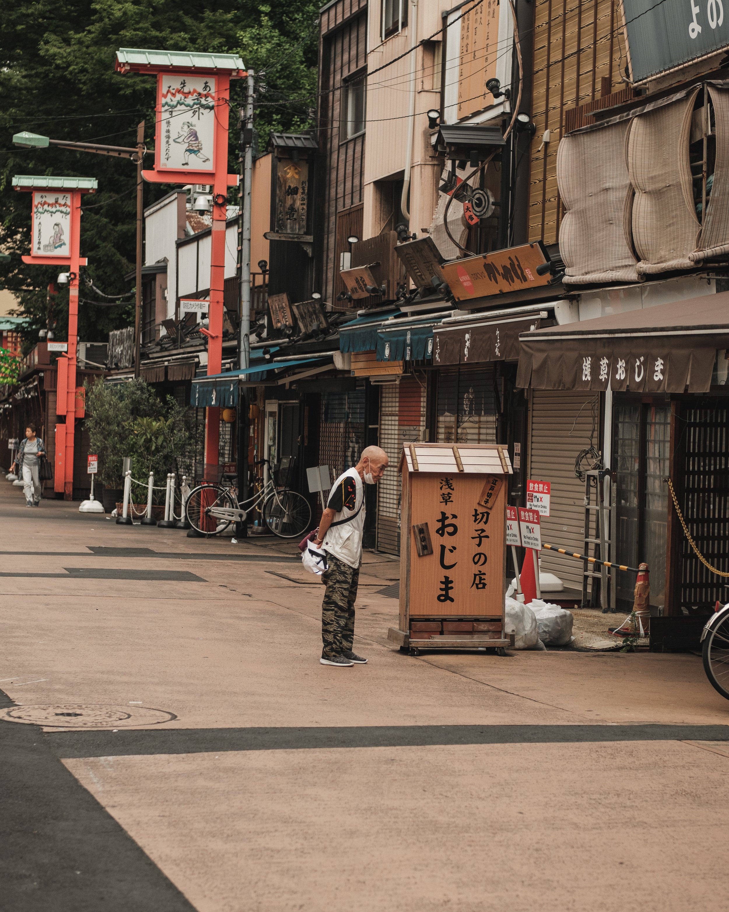 A man wearing a white mask and a vest standing on the sidewalk reading a wooden signboard written in Japanese, with closed shops and bicycles in the background along a street in Japan.