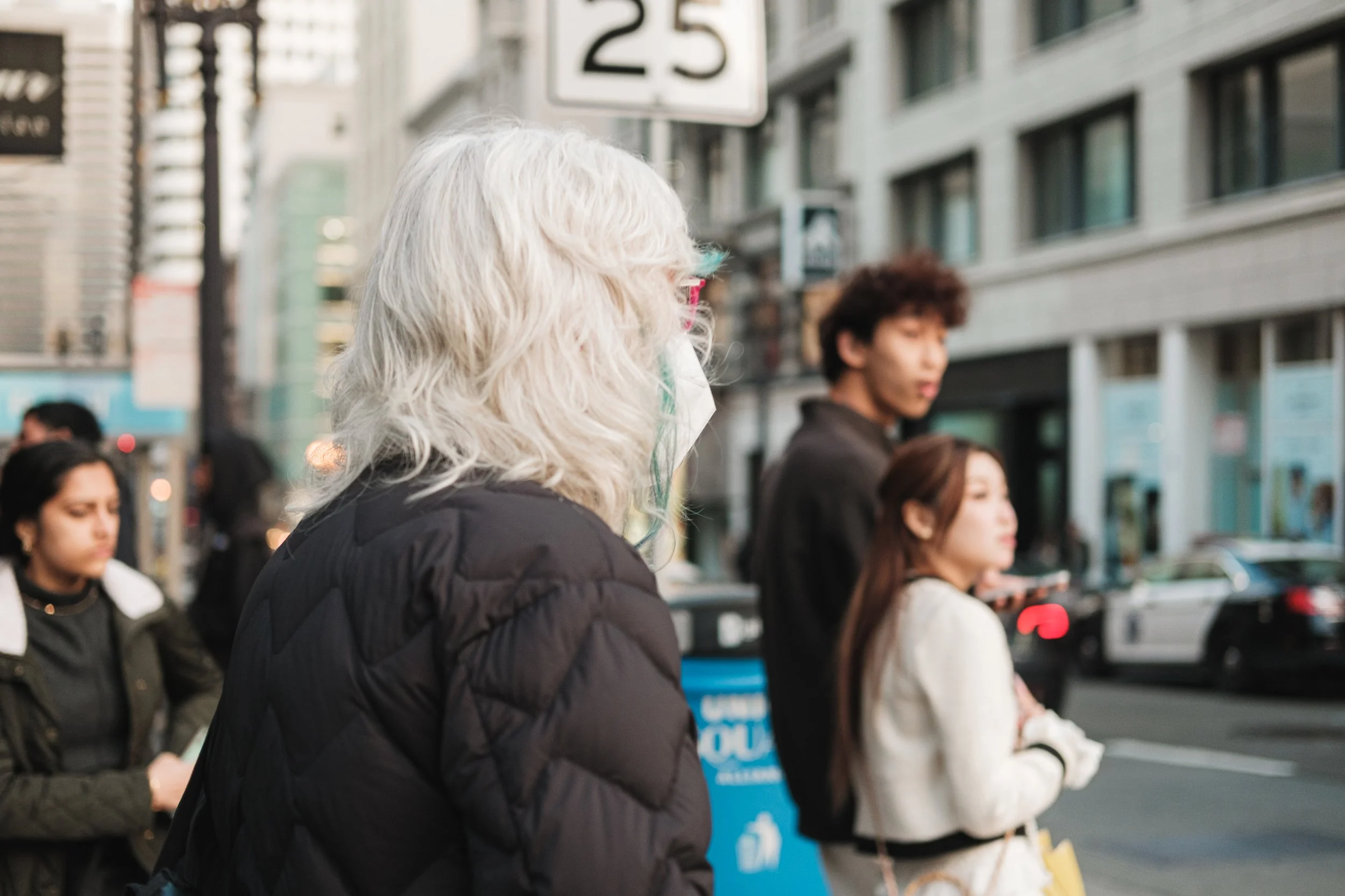 A woman with curly platinum blonde hair wearing a black jacket and a face mask stands on a city sidewalk, with several pedestrians and buildings in the background.