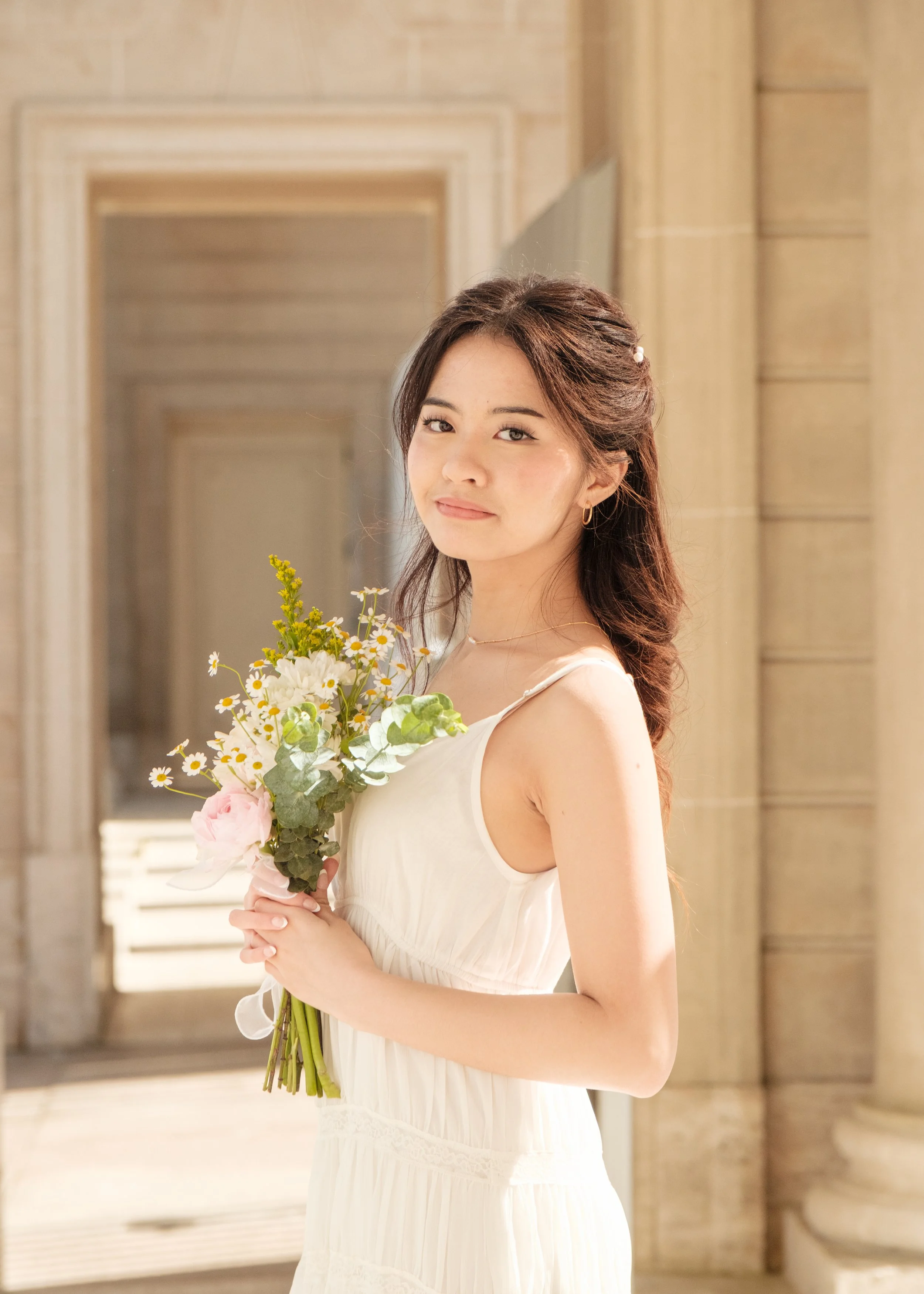 Young woman in white dress holding a bouquet of flowers, standing in a sunlit indoor corridor with classical architecture.