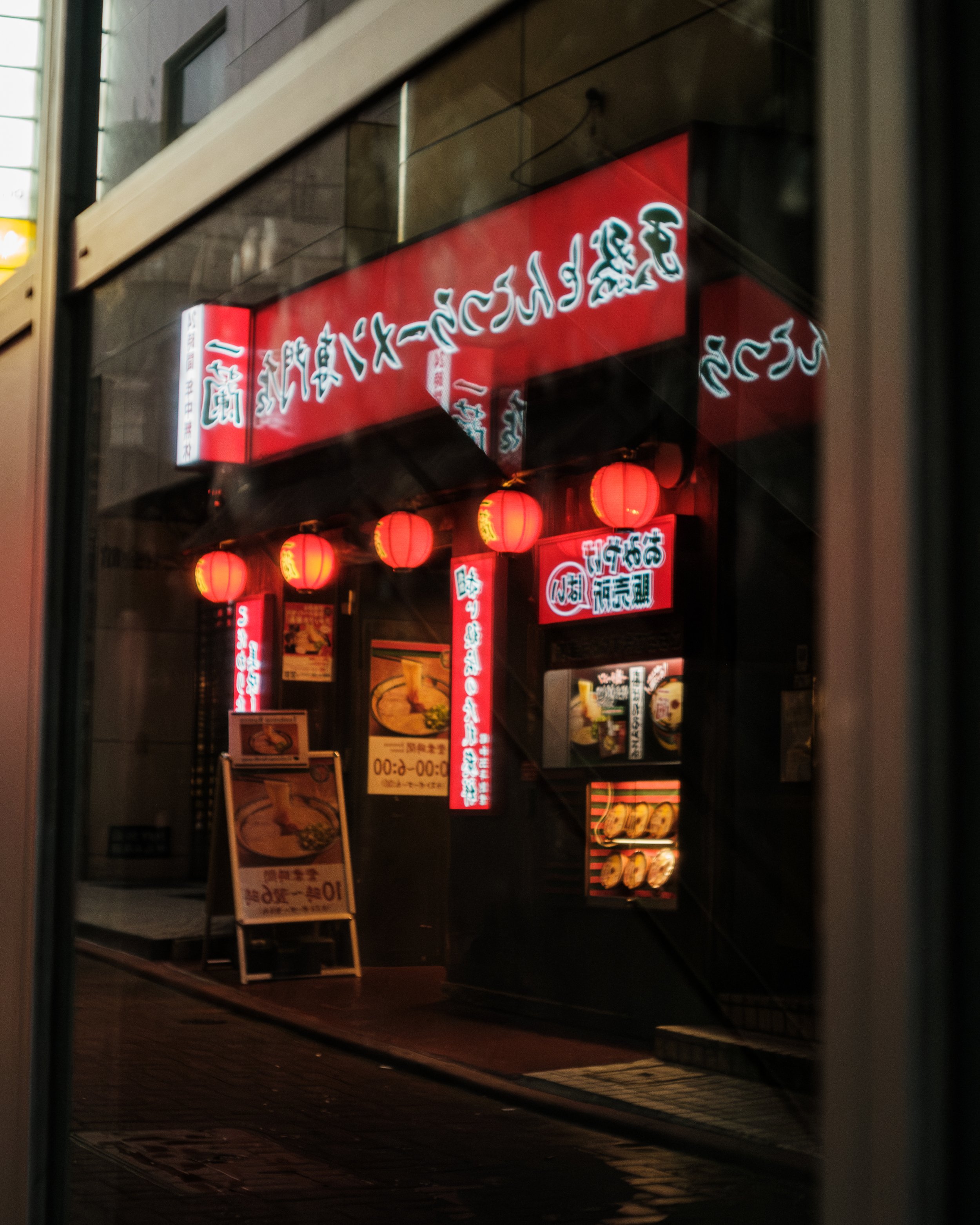 View of a Japanese restaurant illuminated with red neon signs and paper lanterns, seen through a glass window at night.