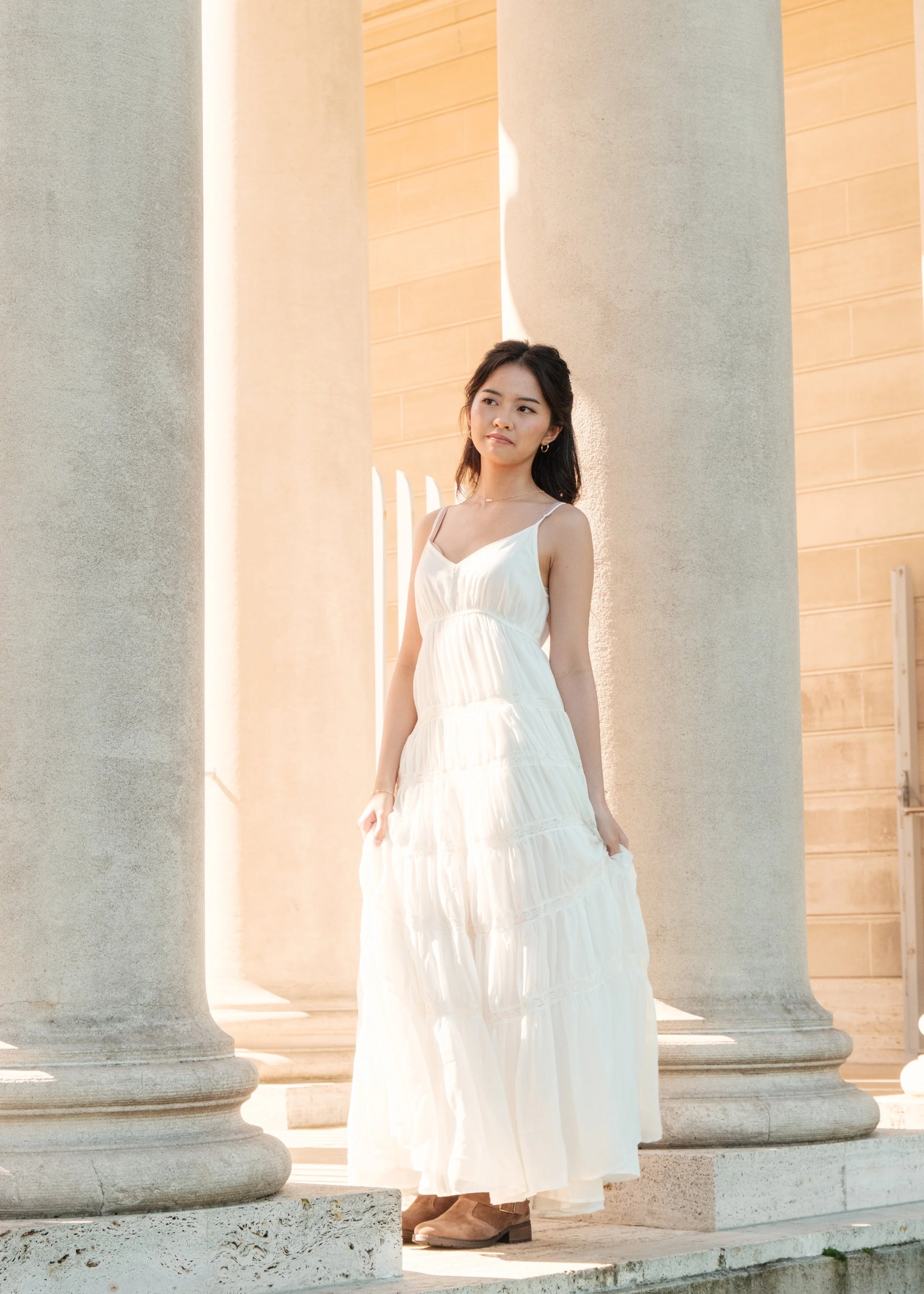 A young woman in a white flowy dress standing between large stone columns in sunlight.