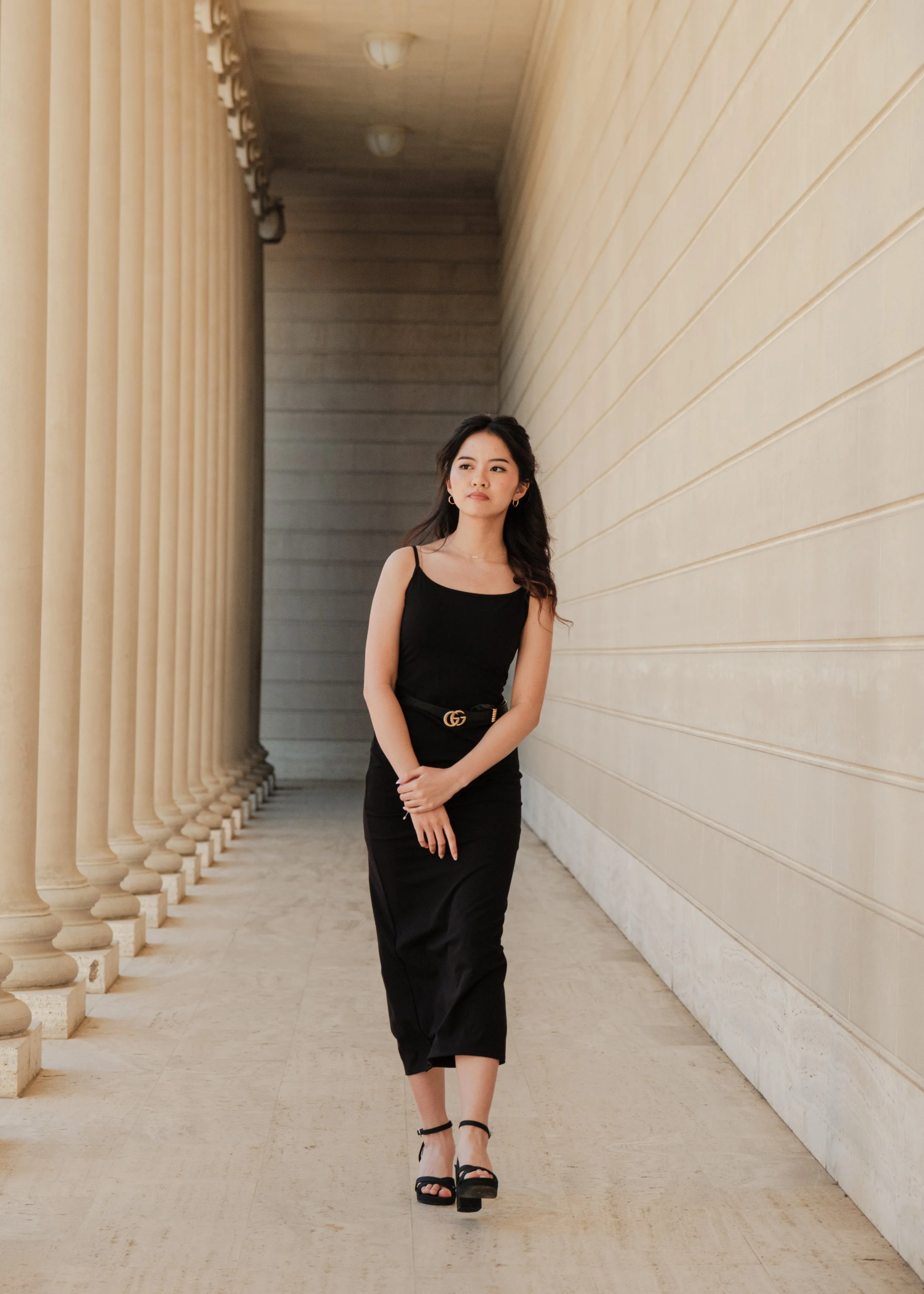 A young woman in a black dress and high heels walking along a walkway with beige columns and a beige wall.