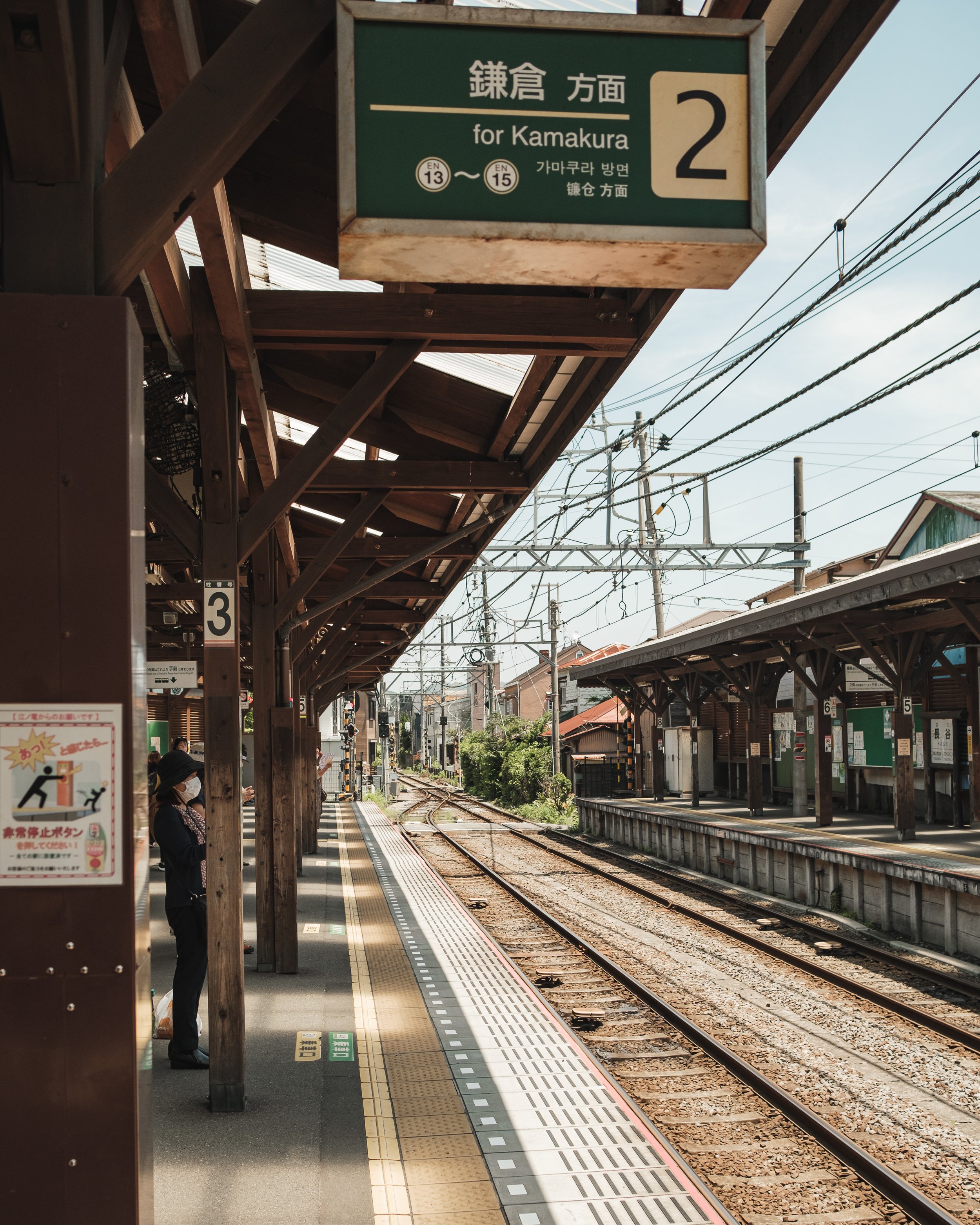 A train station platform with a green sign overhead indicating it is for Kamakura, with platform numbers 2 and 3 visible. There are a few people waiting, wearing masks, under a wooden roof. Tracks run through the station, with overhead electric lines