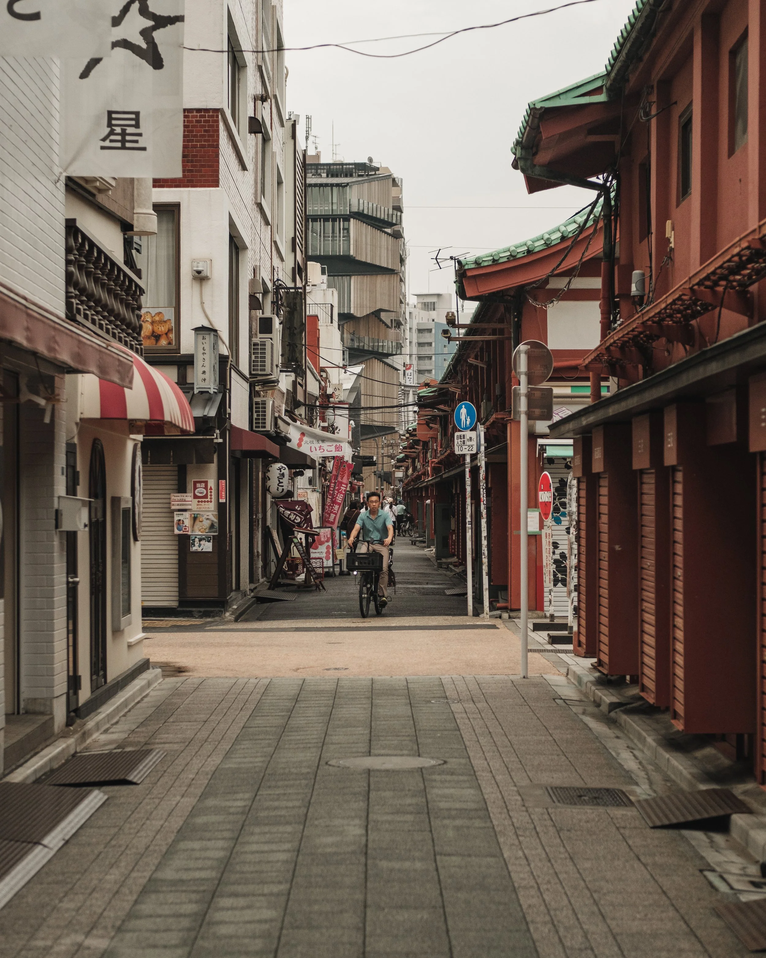 A pedestrian street in Japan with small shops, traditional buildings with red and green roofs, and a man riding a bicycle down the street.