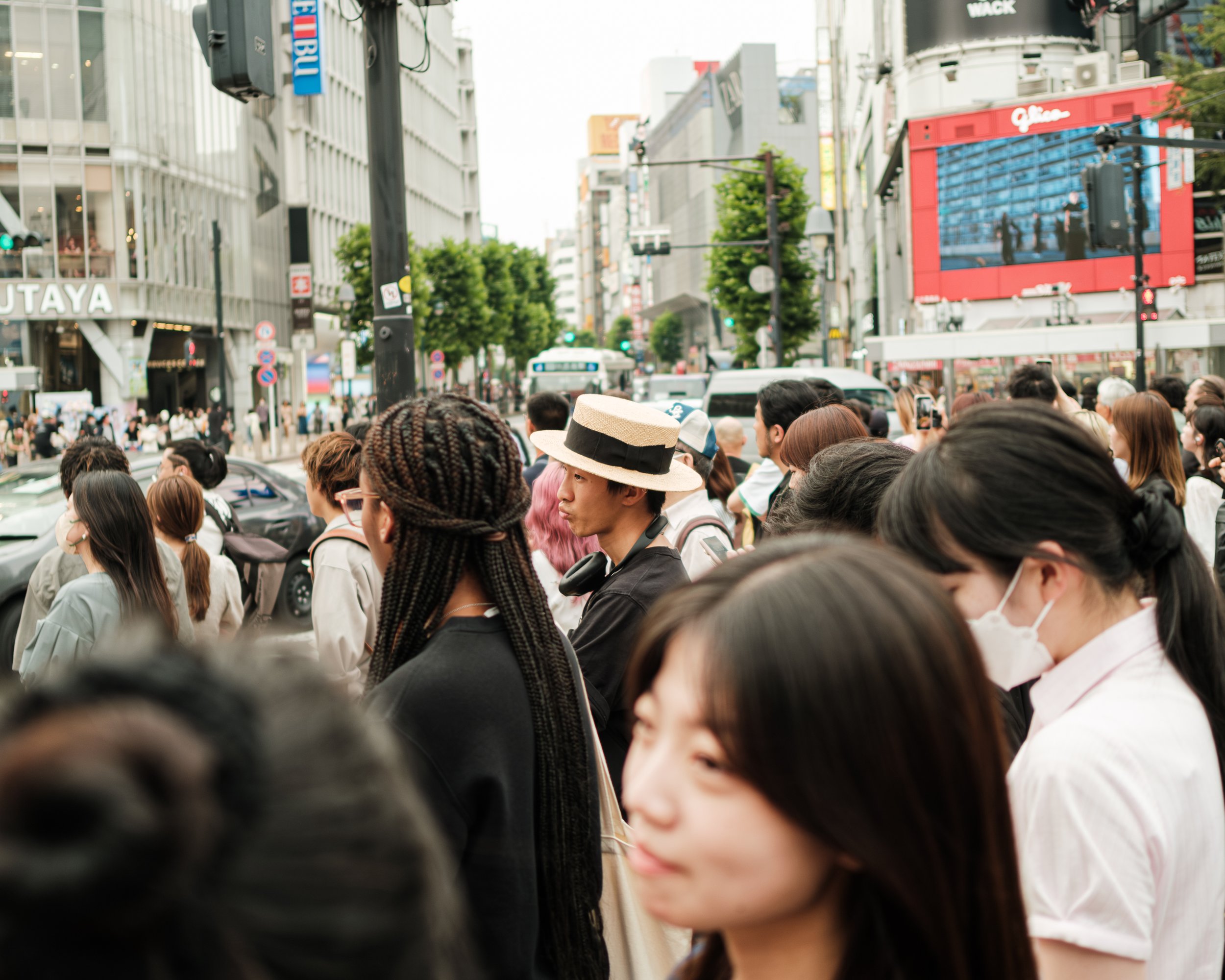 Crowd of people waiting to cross a busy city street on a sunny day, with tall buildings and electronic billboards in the background.