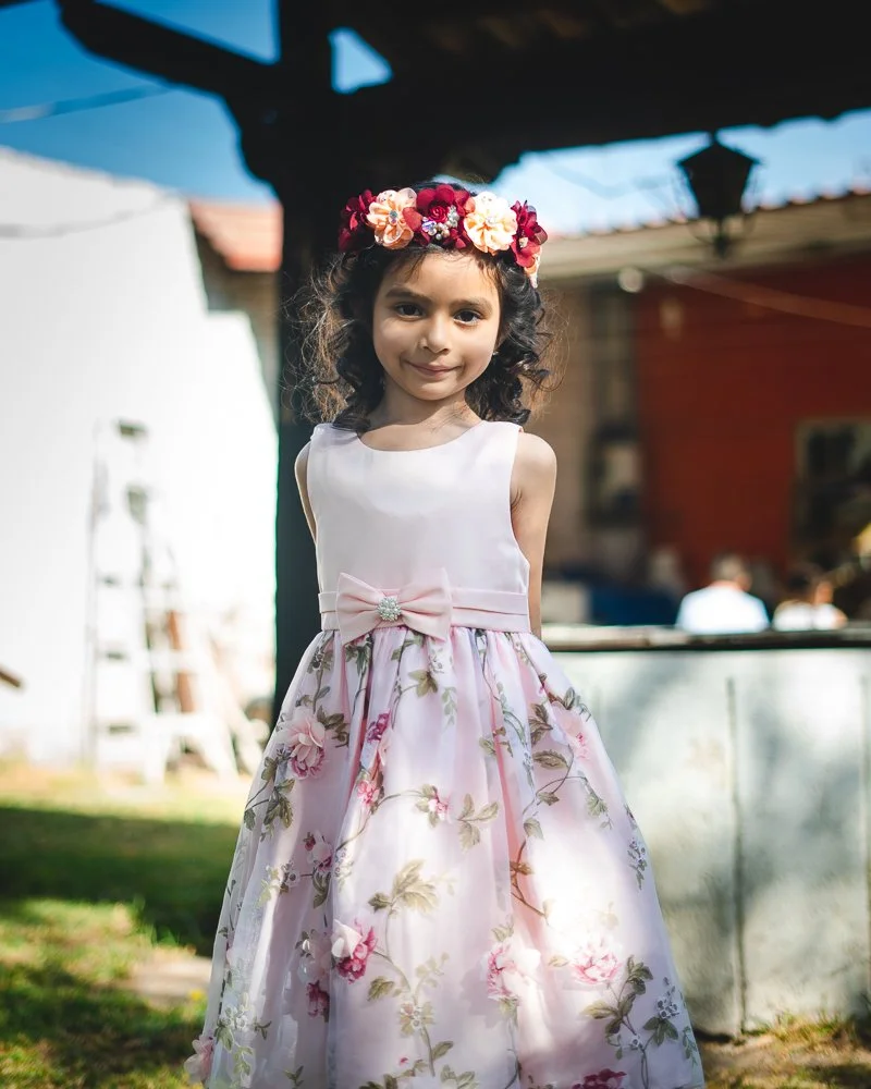 A young girl with dark, curly hair wearing a sleeveless, white and pink floral dress with a pink bow and flower embellishment at the waist, and a flower crown on her head, standing outdoors in sunlight with a blurred background of a white building, t