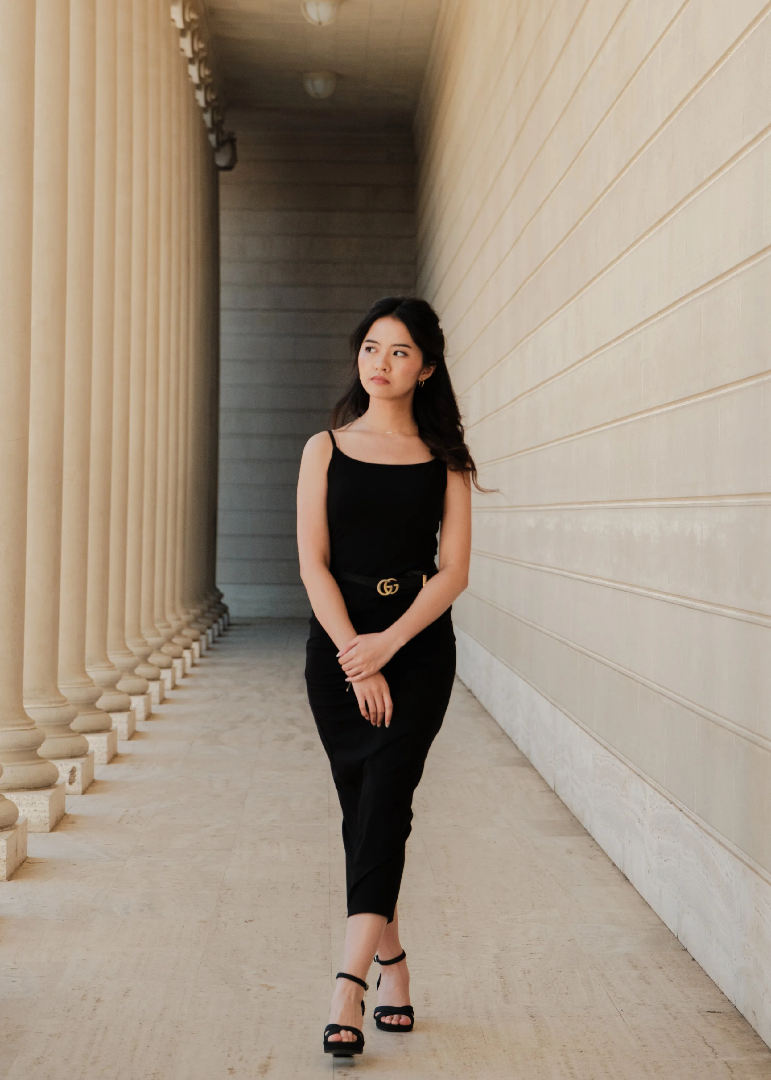 A woman in a black dress and high heels walking along a colonnade with beige columns and paneling.