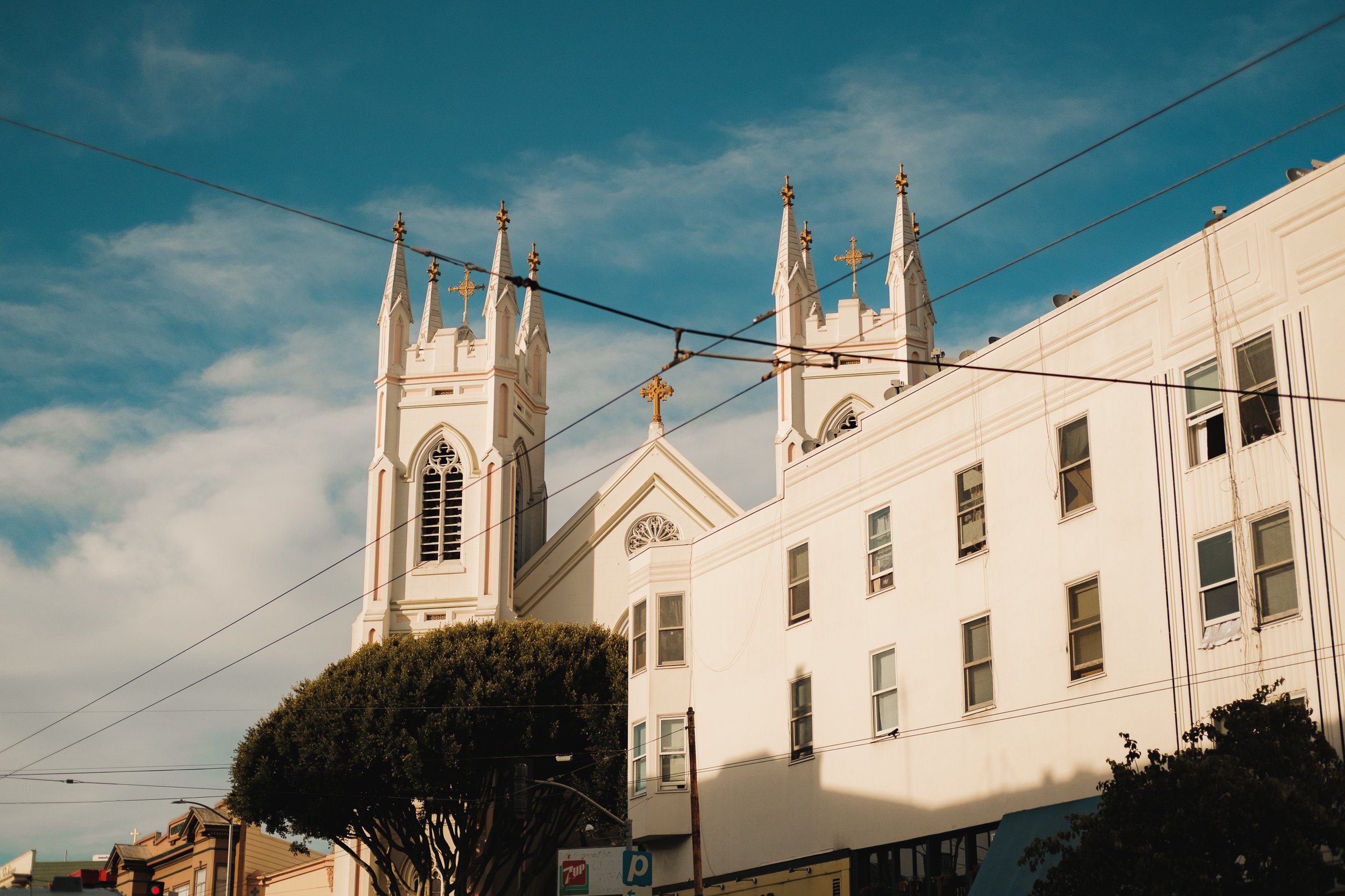 A white Gothic-style church with two tall steeples and a cross on top, set against a clear blue sky with some clouds.