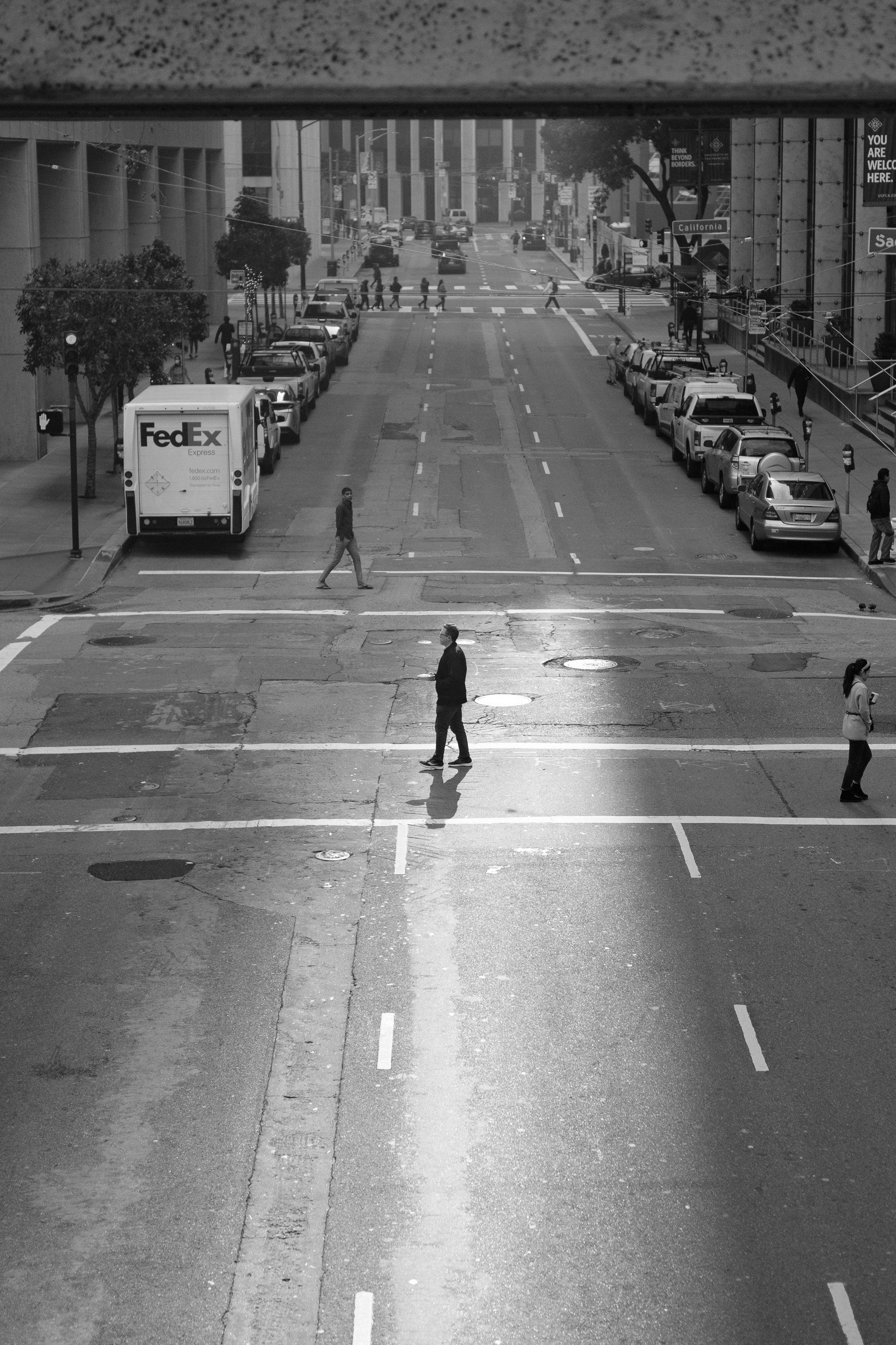 Black and white photo of a city street with pedestrians crossing and parked cars, including a FedEx truck, taken from an elevated position.