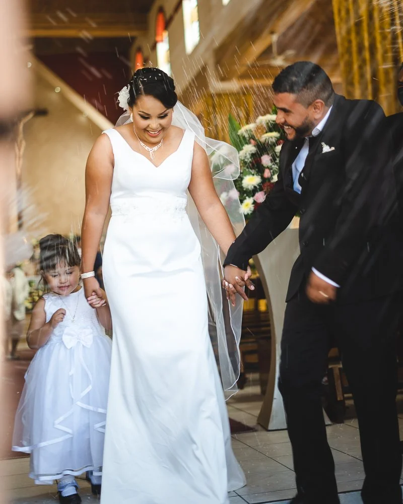 A wedding scene with a bride and groom holding hands, standing together under a shower of rice. The bride is wearing a white wedding dress and veil, and the groom is in a black suit. A young girl in a white dress is holding the bride's hand.