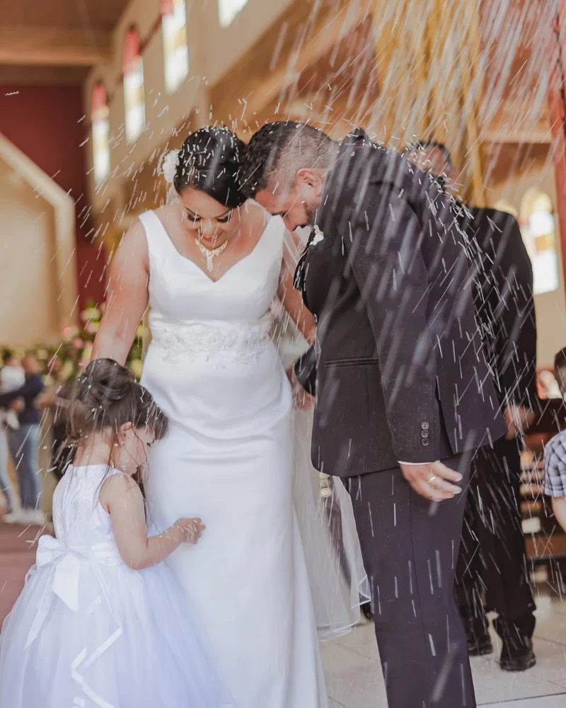 A bride and groom are standing close together, smiling and bowing their heads during a wedding celebration as they are showered with rice or confetti. A young girl in a white dress stands nearby, looking down. The scene is inside a church with wooden