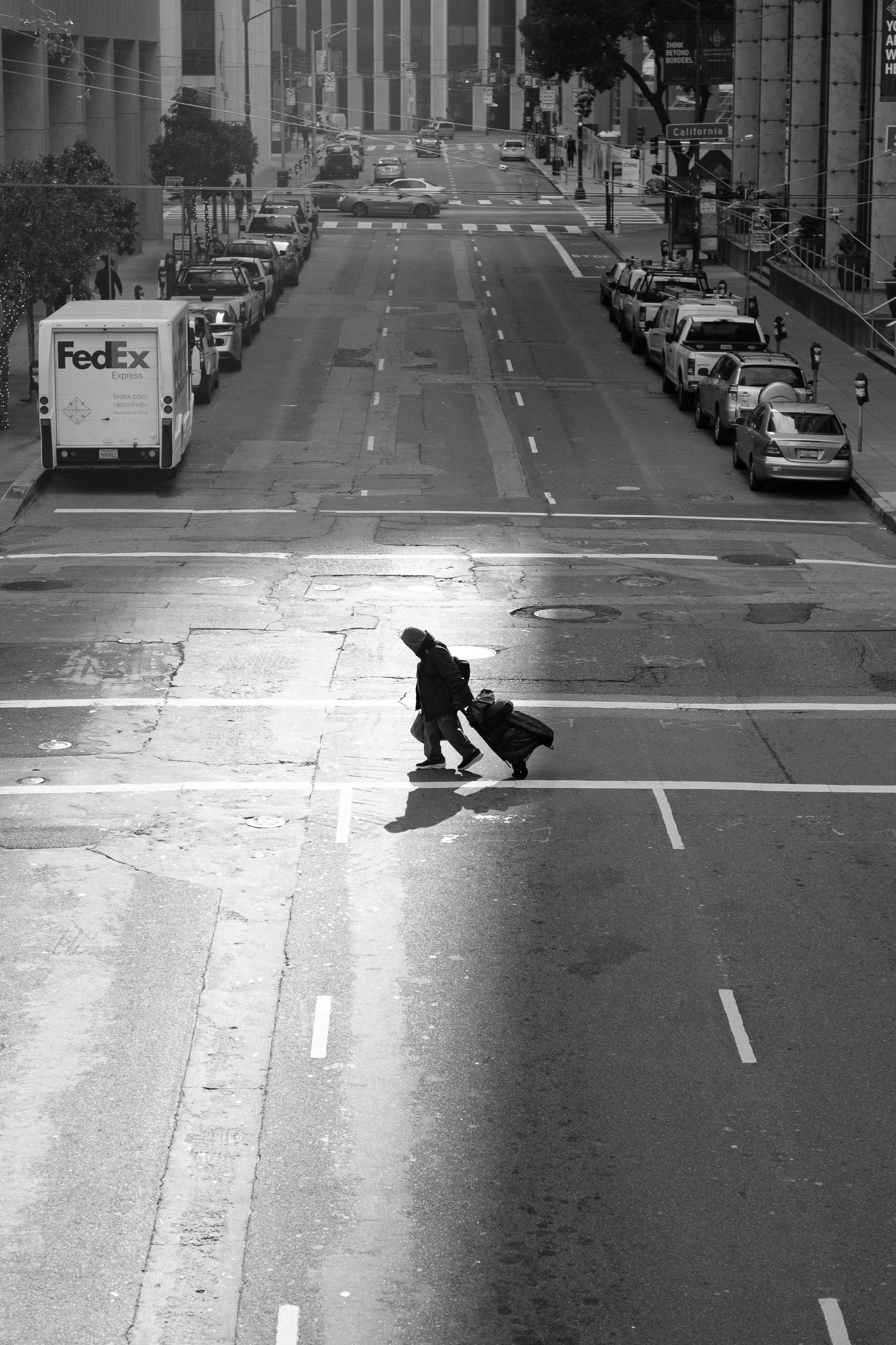 A person with a rolling suitcase crossing an empty city street at sunrise or sunset with parked cars and buildings in the background.