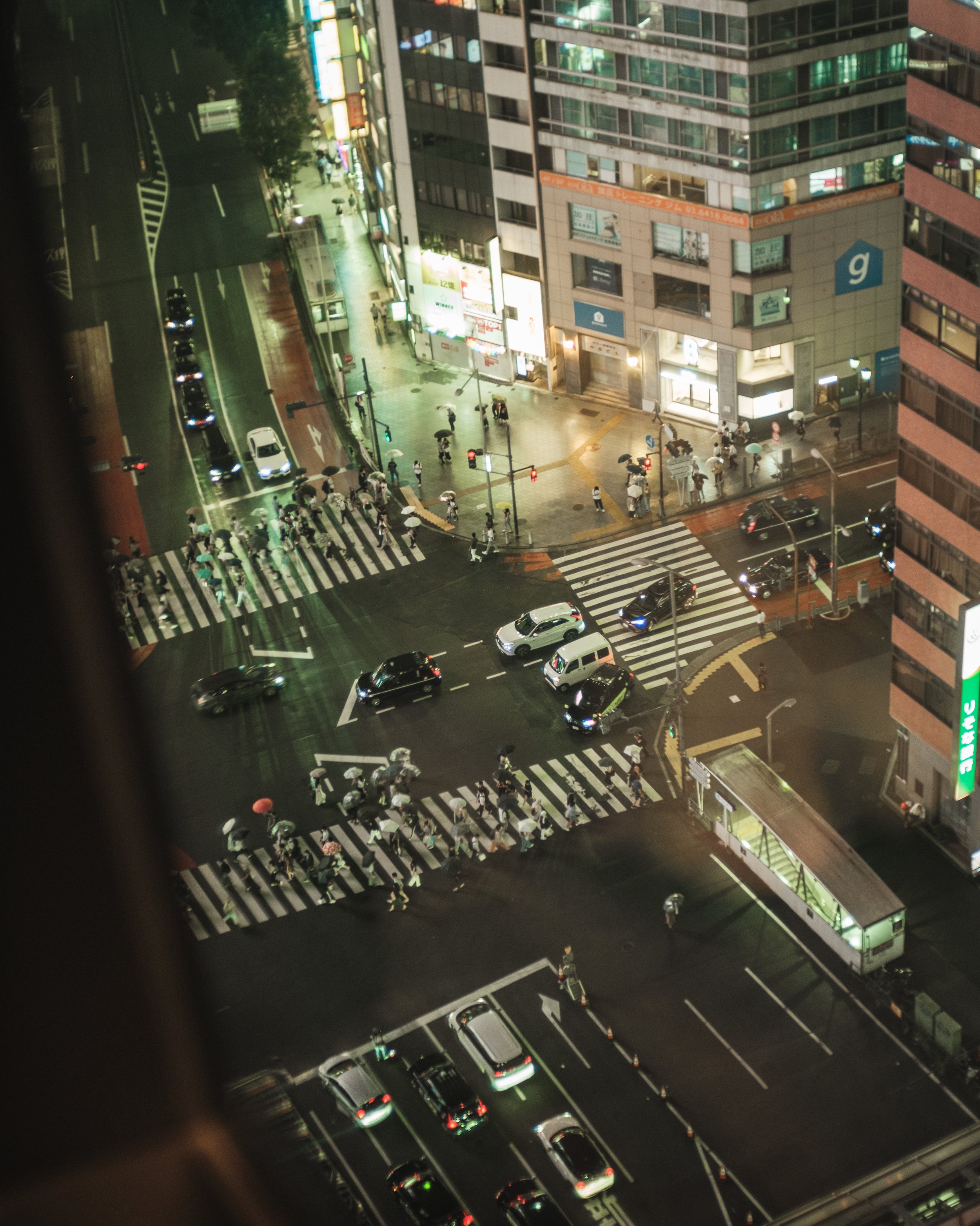 Nighttime city street scene with pedestrians crossing at crosswalks, cars on the road, illuminated buildings, and a parking lot beneath the view.