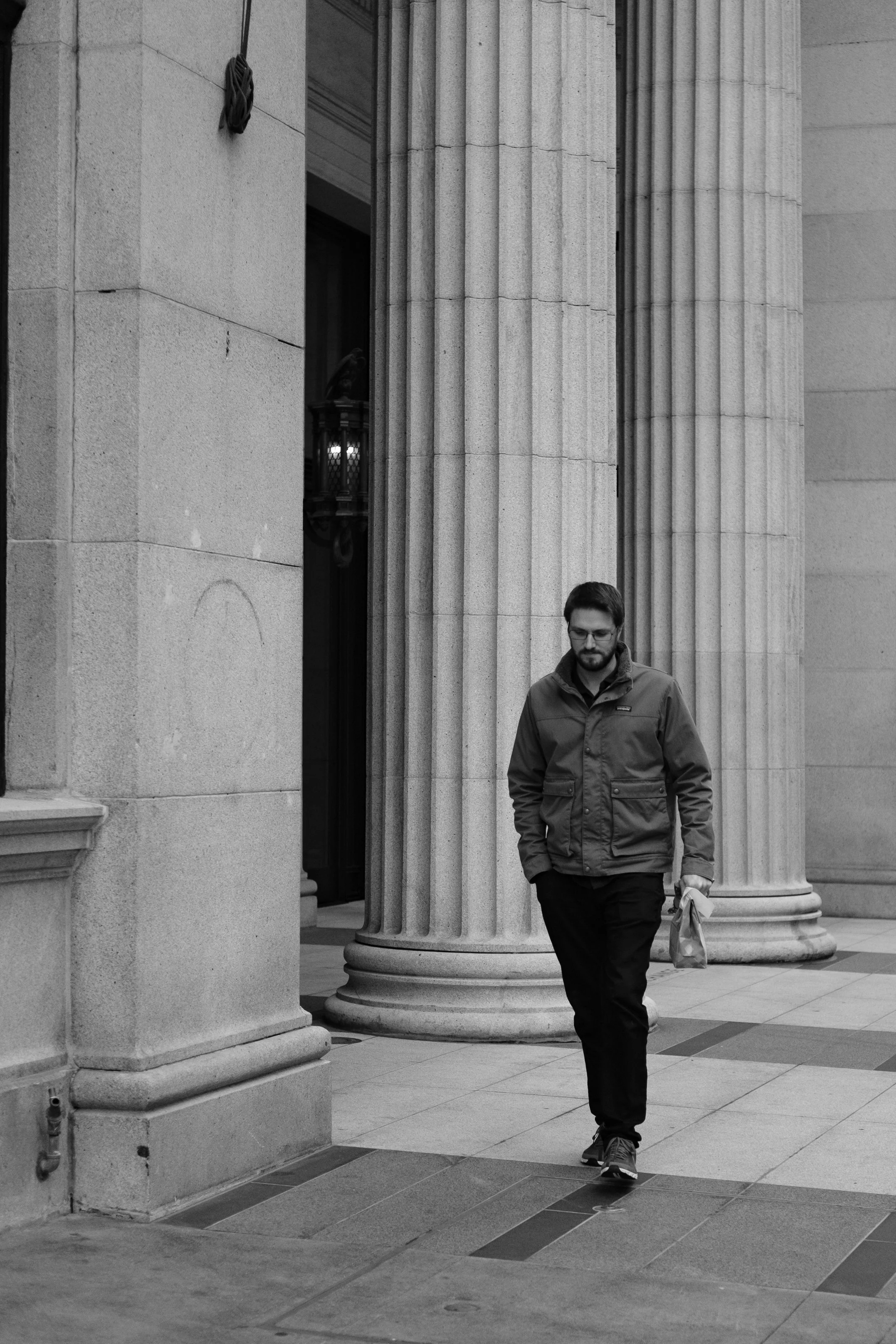 A man walking past large stone columns on a city street, holding a small shopping bag, wearing glasses and a jacket.