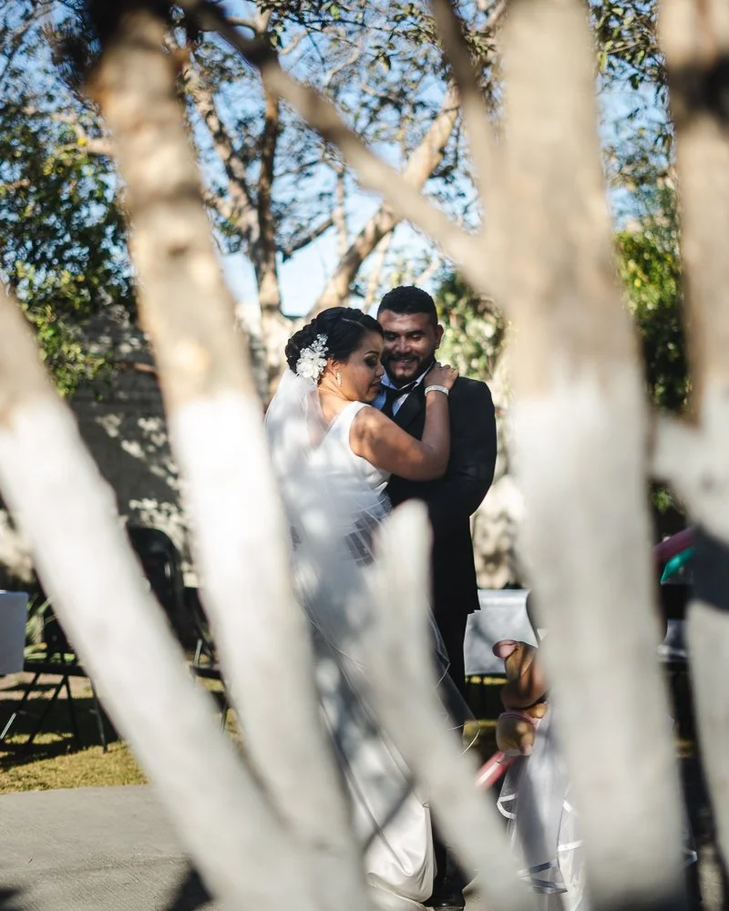 A bride and groom embracing outdoors during their wedding, seen through tree branches.