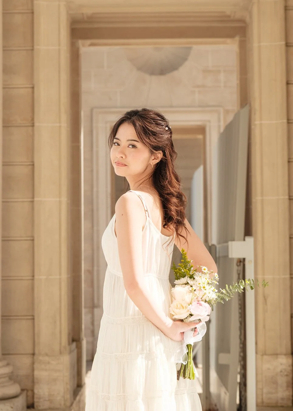 A young woman in a white dress holding a bouquet of white and pink flowers stands in an elegant stone hallway with archways.