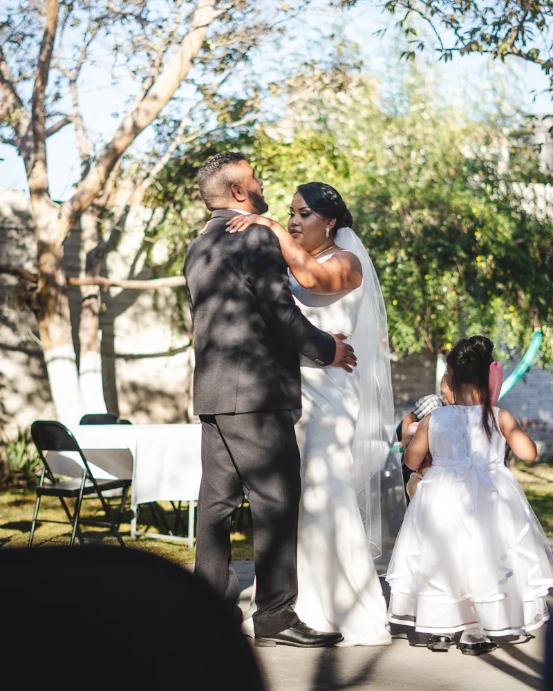 A bride and groom dancing outdoors at their wedding, with a young girl in a white dress with a pink bow watching them.