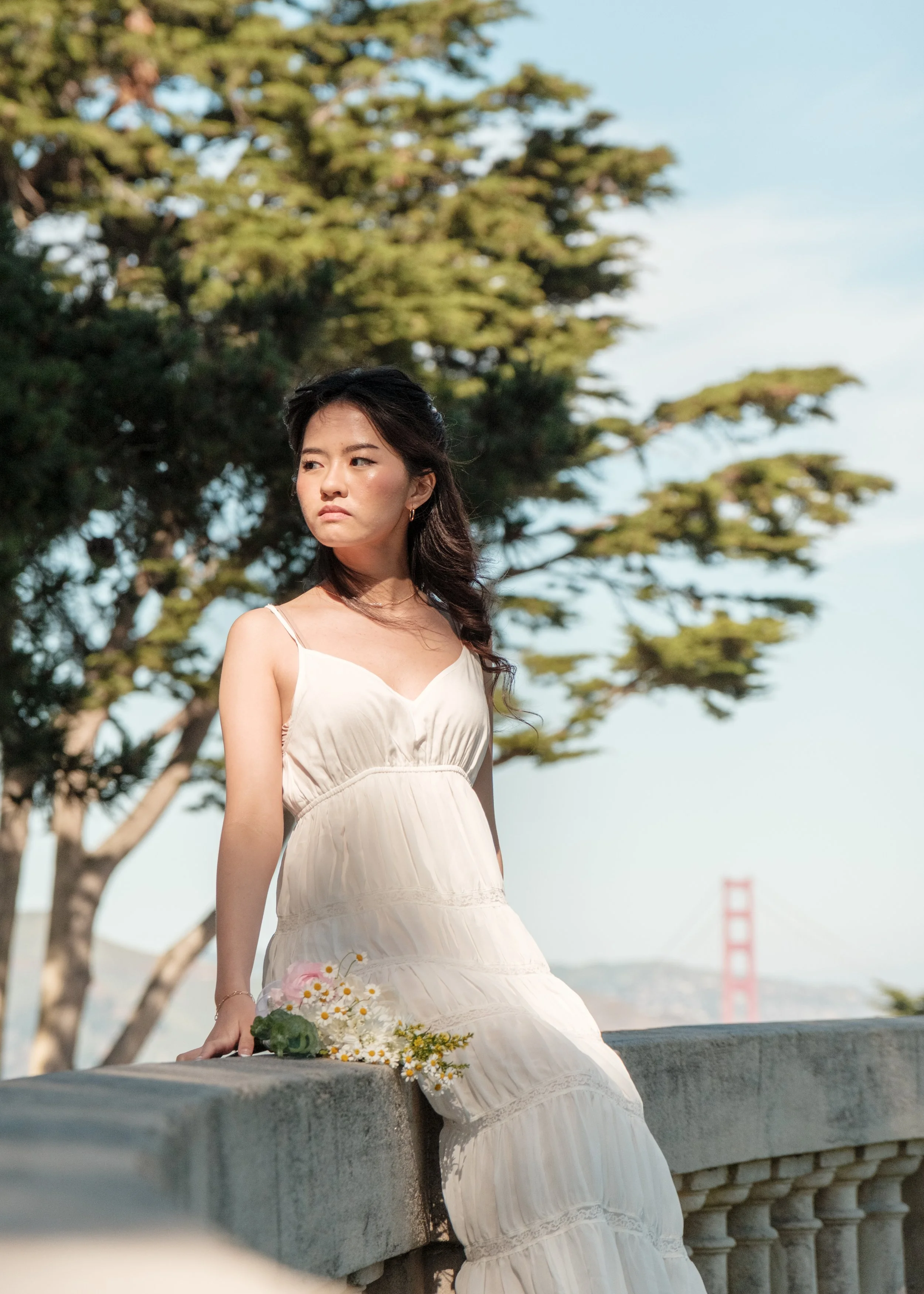 A woman in a white summer dress sitting on a stone ledge with flowers, trees, and the Golden Gate Bridge in the background.