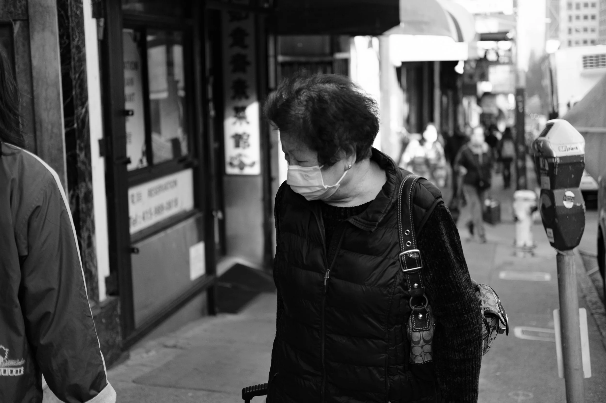 A woman wearing a face mask, a black vest, and carrying a purse, standing on a city sidewalk in front of a storefront with Chinese characters on the window.
