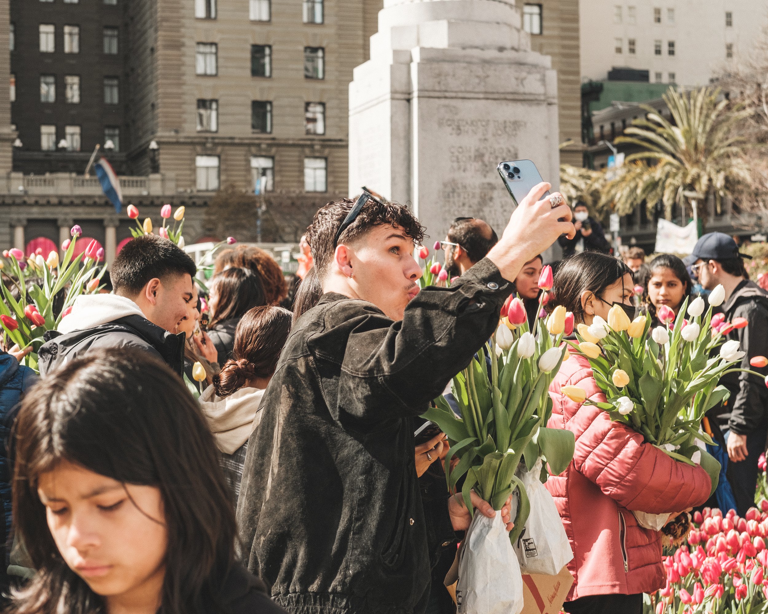People gathering outdoors in a city park with tulips, with some taking photos and holding flowers, during a sunny day.