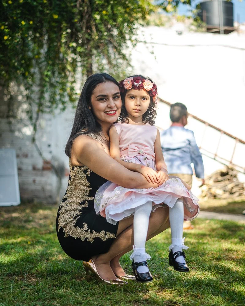 A woman in a black and gold embroidered dress holding a young girl in a pink dress and floral headband, outdoors on grass.