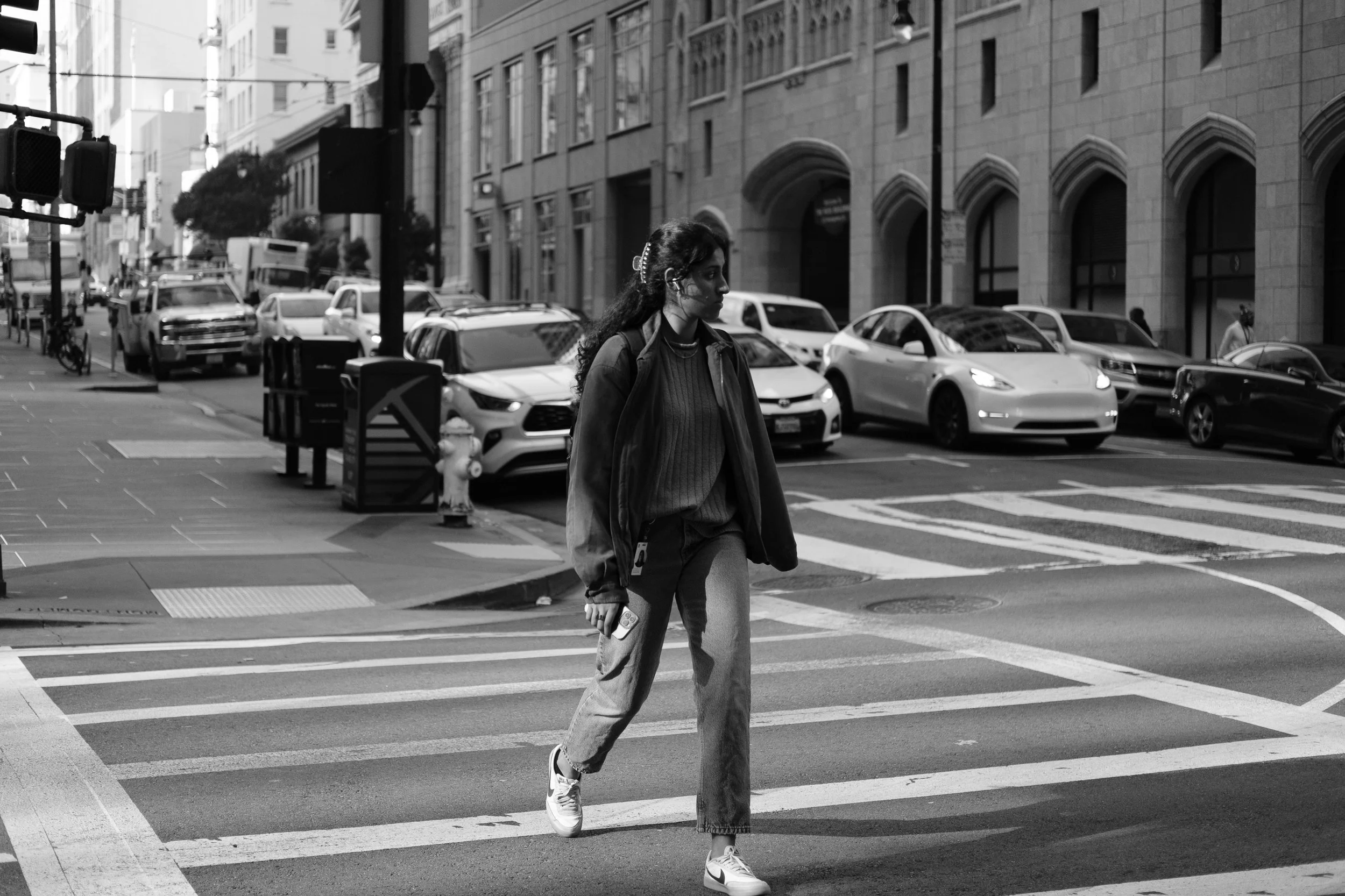 A woman walking across a city street in black and white, carrying a backpack and walking on a crosswalk with parked cars and buildings in the background.