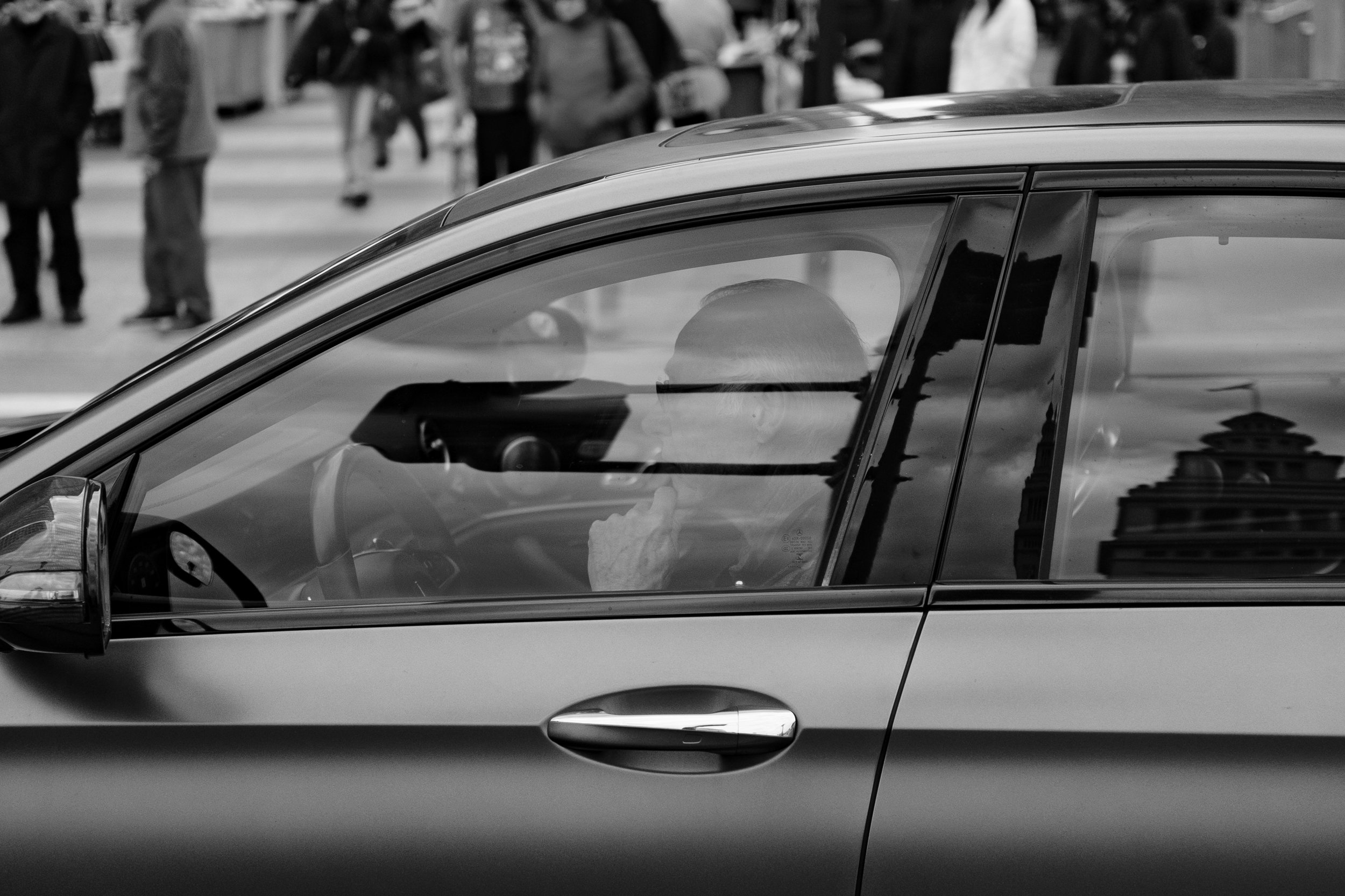 A man seated inside a car, looking at his phone, with a blurred city street and pedestrians in the background.