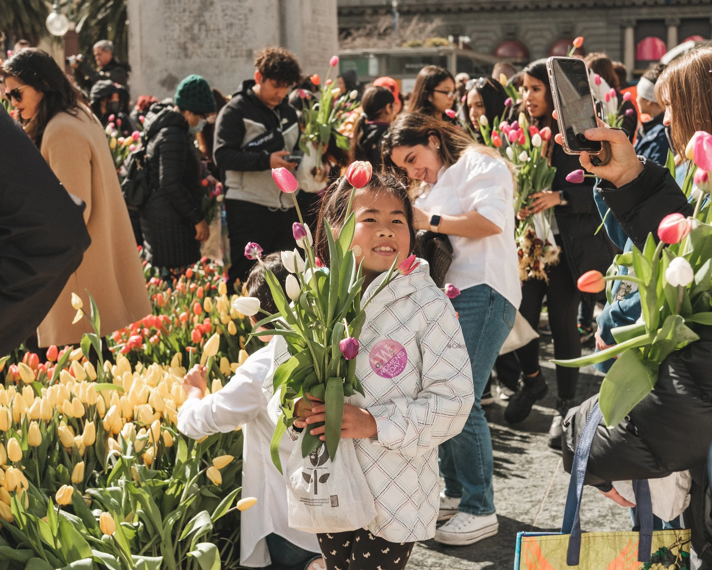 A girl holding a bouquet of tulips at a flower market, surrounded by people also holding flowers and taking photos of the flowers.