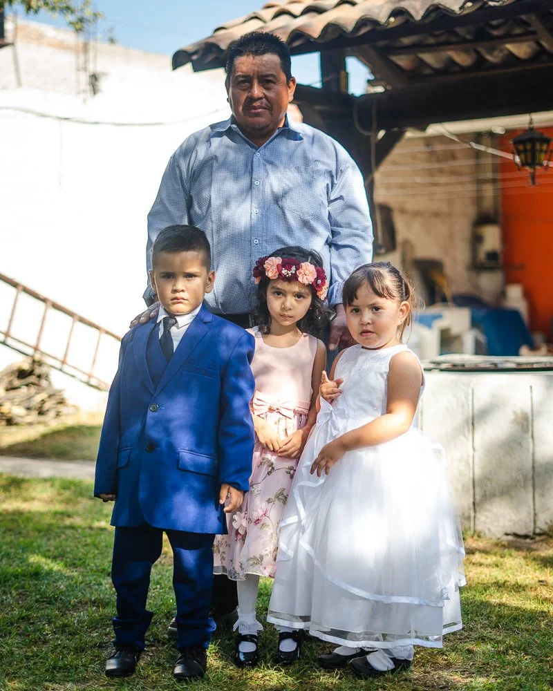 A man and three young children posing outdoors. The man is standing behind the children, wearing a light blue button-up shirt. The children are dressed in formal attire: a boy in a blue suit and tie, and two girls in white dresses, one with a floral 