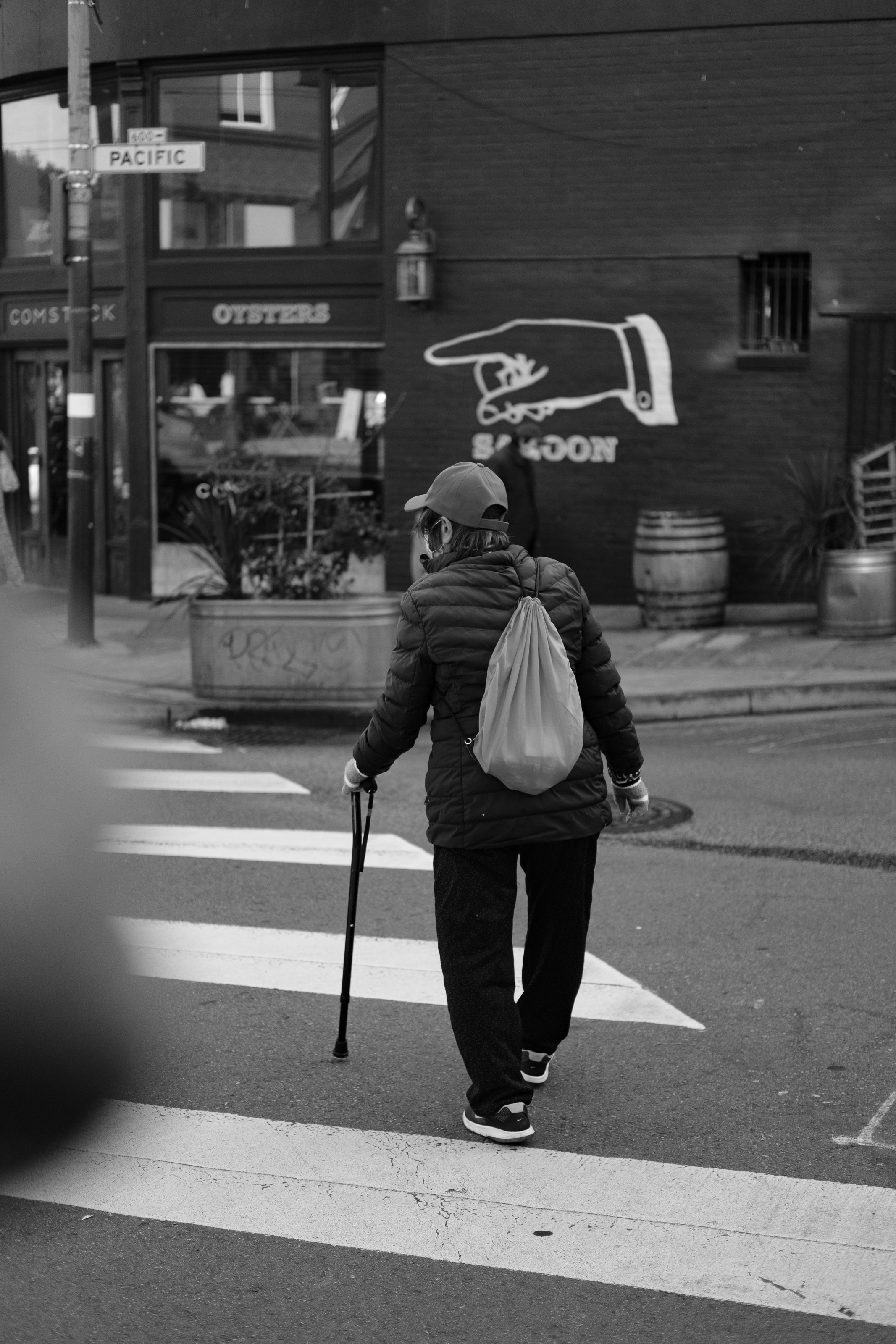 A woman walking across a crosswalk with a cane, wearing a backpack and cap, in an urban setting with storefronts and street signs in the background.