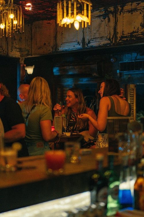 A group of people socializing in a dimly lit bar, with drinks on the counter and warm ambient lighting.